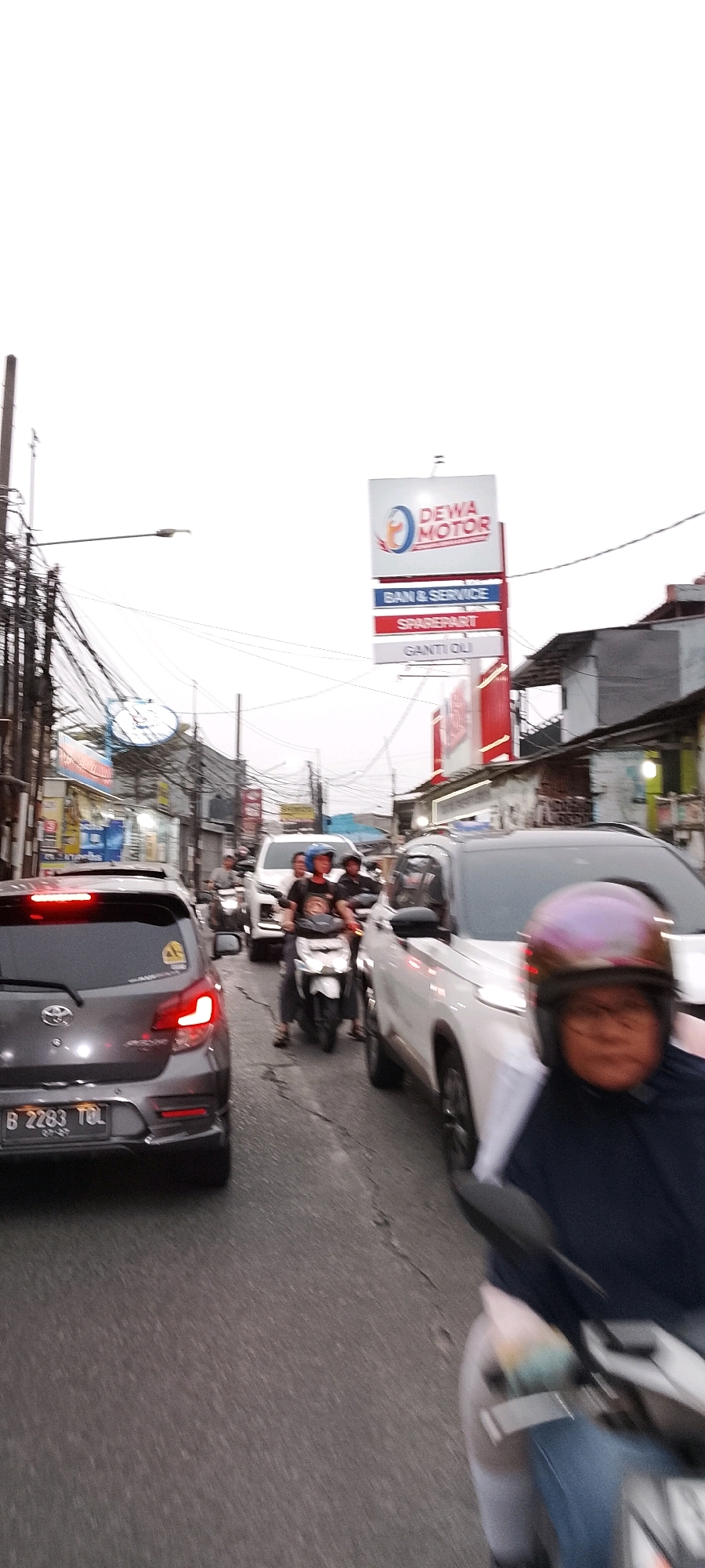 The image depicts a street scene in Depok, Indonesia, likely during daylight with an overcast sky. The foreground is dominated by the rear of a dark grey hatchback car with its brake lights illuminated and a license plate reading "B 2283 TOL". To the right, a white SUV is partially visible. The middle ground shows several motorcycles and cars in traffic. One motorcycle in the center has two riders; the front rider wears a blue helmet and a black t-shirt with a graphic, while the rear rider is obscured. Another motorcycle further back has a rider with a helmet. In the background, buildings line the street, and overhead wires are prominent. A large sign with "DEWA MOTOR" and services like "BAN & SERVICE," "SPAREPART," and "GANTI OLI" is visible, indicating a motor vehicle service center. The overall environment suggests a busy urban street with moderate traffic.
