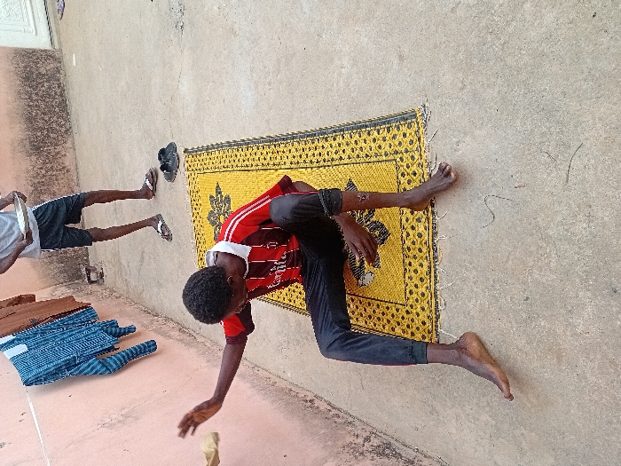 An outdoor scene in Ayama, Nigeria, features two males on a concrete surface against a light pink wall. In the foreground, a dark-complexioned male sits barefoot on a yellow woven mat with black geometric patterns. He wears a red and black striped short-sleeve shirt and black pants, with an apparent mark or injury on his right lower leg. His right arm is raised, hand open, seemingly interacting with a light-colored, indistinct object held near his hand. Behind him, to the right, a second dark-complexioned male stands, wearing a light grey t-shirt, dark shorts with white side stripes, and white flip-flops. He holds a light-colored shallow bowl or plate in both hands. Near his feet on the concrete are two dark shoes or slippers. On the pink wall to the left, a blue and white patterned long-sleeve shirt and a brown garment hang. A white door is partially visible in the far background. The ground is concrete with minor debris.