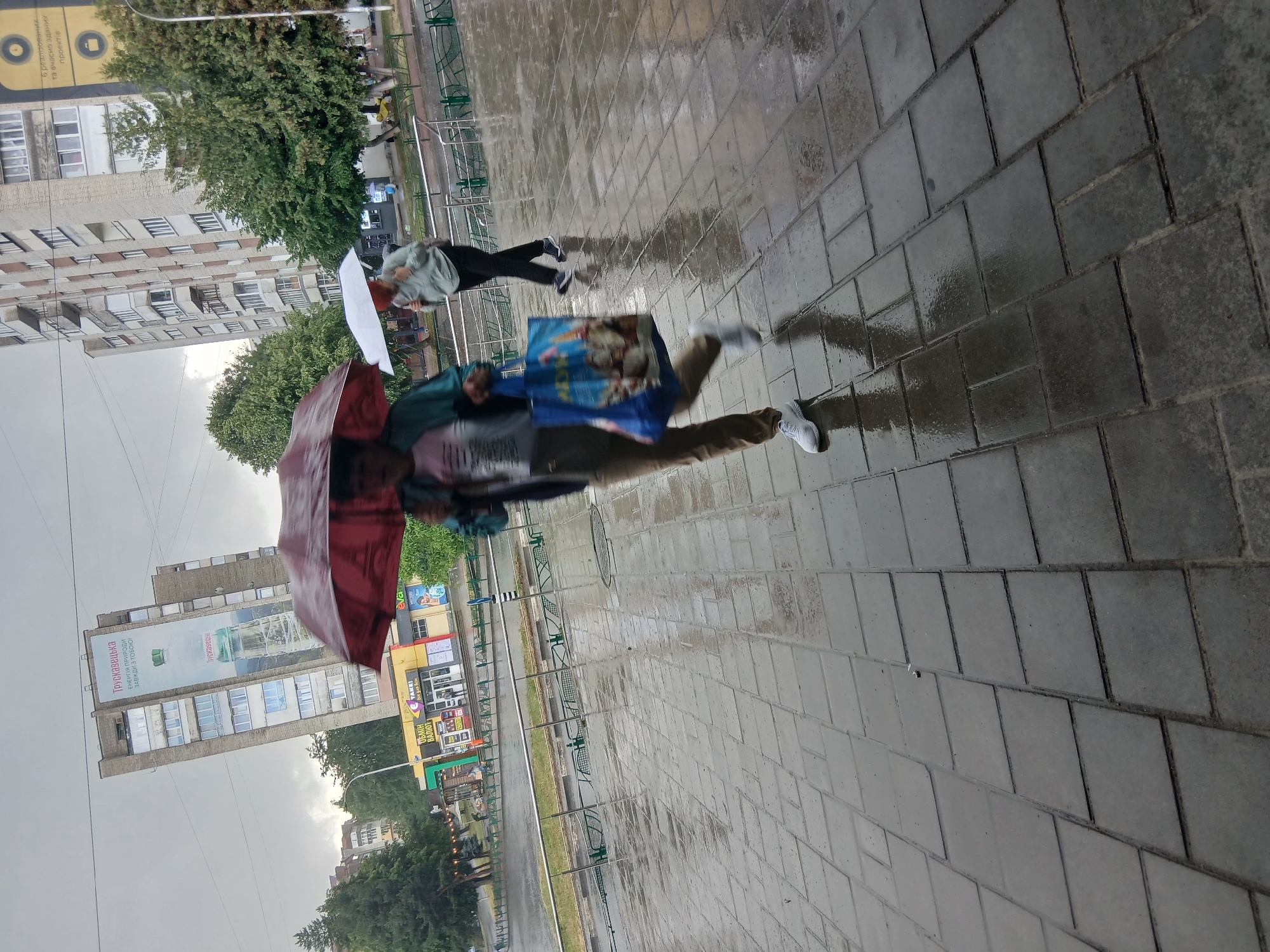 In Truskavets, Ukraine, on a cloudy, wet day, two individuals are seen walking under umbrellas on a wet, tiled pedestrian walkway. The foreground is dominated by the wet paving stones, reflecting the overcast sky. To the left, a person holds a dark red umbrella and carries a large blue shopping bag with a cartoon image. They are wearing a light-colored t-shirt, a dark jacket, and khaki pants. To the right, another person walks with a white umbrella, wearing a light-colored hooded jacket and dark pants. In the background, a multi-story building with a large advertisement billboard is visible, along with trees and other urban structures. The scene suggests a typical rainy day in a city environment.