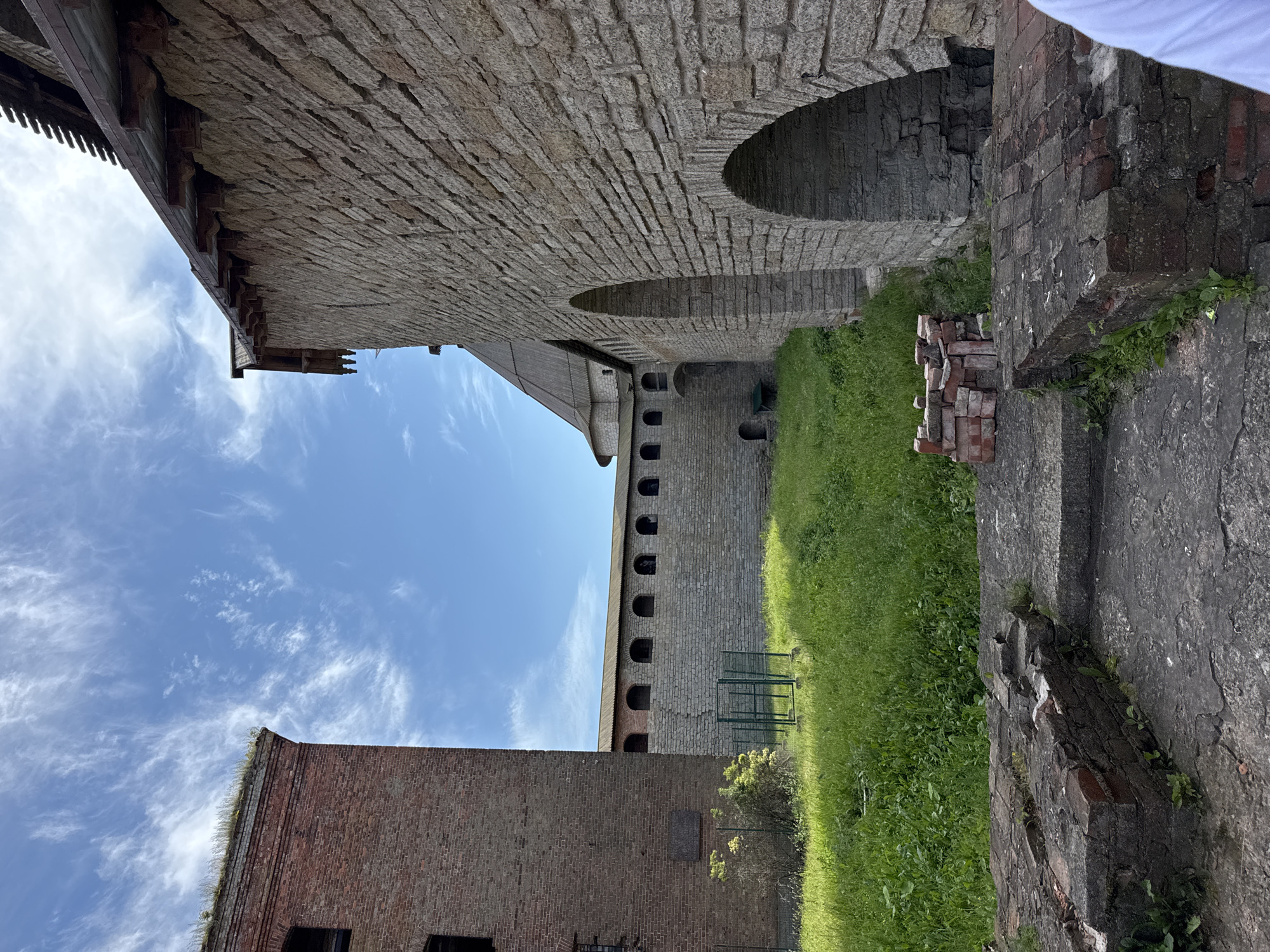 This is a landscape photograph taken from a low angle, featuring an outdoor view of an old stone building with a brick structure in the foreground. The image is rotated 90 degrees counterclockwise, making the sky appear on the left and the ground on the right.
The primary subject is the weathered stone wall of a historical building, constructed with large, irregular stone blocks. The wall features arched openings, suggesting a courtyard or passage. Below this, a long, narrow building with multiple small arched windows is visible, also constructed of stone. In the foreground, a red brick building stands, appearing older and more dilapidated than the stone structure. Piles of red bricks are scattered near this building, alongside overgrown green grass. A small green metal fence is also visible near the stone building. The sky above is blue with wispy white clouds.
The scene depicts an outdoor architectural setting, likely a historical fortress or old industrial complex, given the robust stone construction and the presence of brickwork. The lighting suggests it is daytime, possibly late morning or afternoon, with clear weather. The overall impression is one of historical significance and perhaps a sense of abandonment or disrepair due to the state of the brick building and scattered materials. There is no visible text in the image. The location context provided is Shlisselburg, Russia.