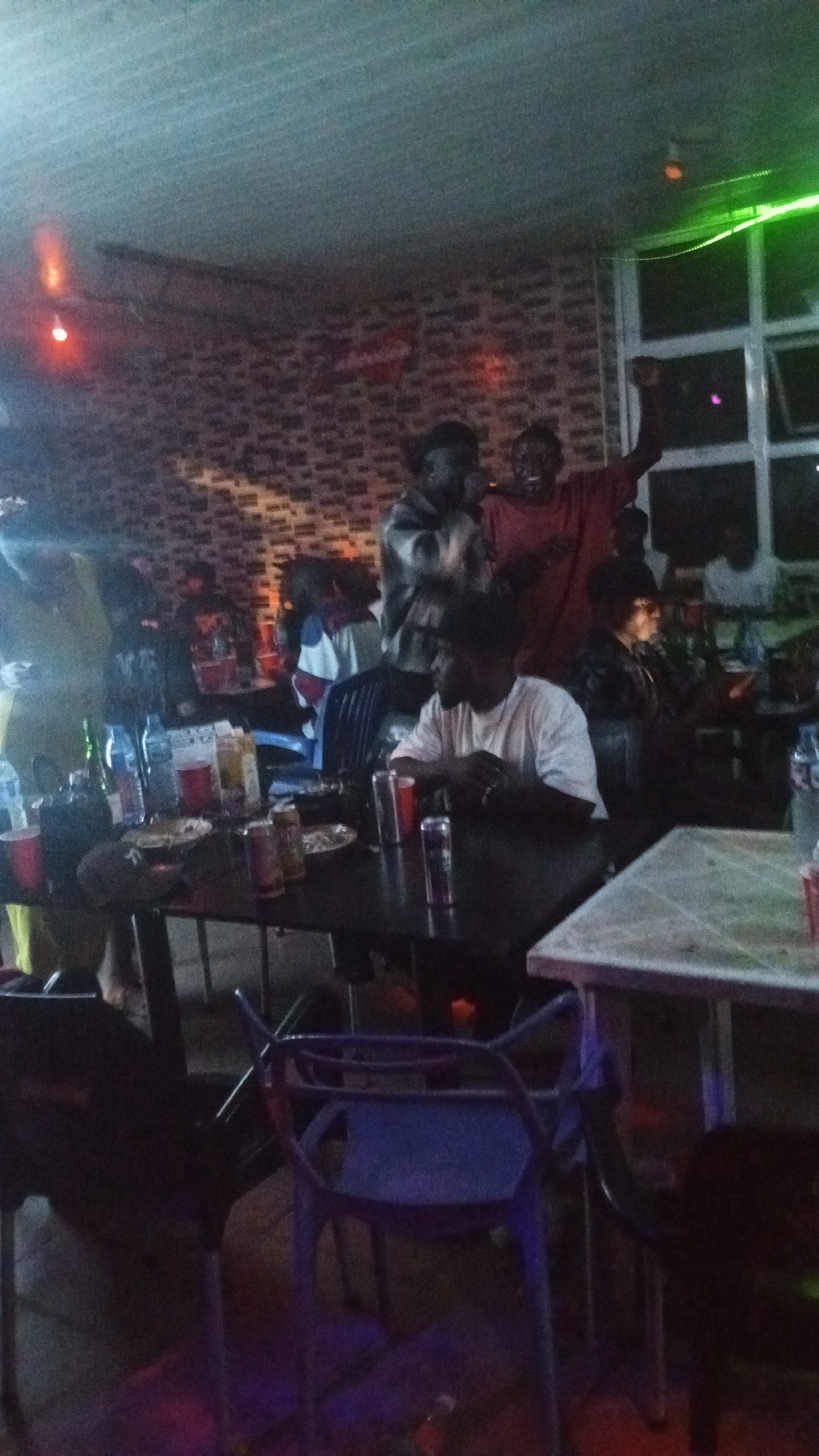 The image depicts an indoor gathering, possibly a bar or club, in an urban setting. The scene is dimly lit, with vibrant green neon lights visible in the background, suggesting a lively atmosphere. A group of young people are congregated around tables, some standing and others seated. Several individuals are holding drinks and appear to be engaged in conversation or celebration. The walls are decorated with a patterned wallpaper, and the furniture includes tables and chairs. The overall impression is one of a social event or a night out. The location is identified as Unknown, Nigeria.