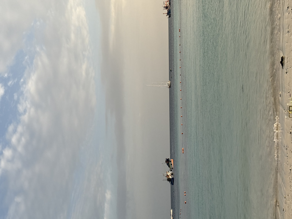 The image depicts a tranquil coastal scene on Ban Ko Li Pe, Thailand, viewed from a sandy beach towards the open sea under a partly cloudy sky. The sky, occupying the upper third of the frame, features a mix of light blue patches and scattered white and grey clouds, with a subtle warm glow on the right horizon. The middle ground is dominated by calm, clear turquoise water, transitioning to a deeper blue towards the horizon. A string of approximately sixteen evenly spaced orange buoys forms a linear barrier across the water, delineating an area closest to the shore.
Several vessels are visible on the water. To the left, a large, dark-hulled fishing or utility boat with a white superstructure is anchored. Towards the center-right, a white sailboat with two tall masts is also at anchor. On the far right, part of a traditional Thai longtail boat or similar vessel is visible. A small, white powerboat or dinghy is partially visible on the far left.
The foreground consists of light-colored sand, with gentle waves breaking at the water's edge, creating a thin line of white foam. Small pieces of natural debris, including a dark mass, are present on the wet sand near the bottom center. A small, circular metallic object, possibly a ring or part of a structure, is partially buried in the sand in the lower central portion of the image. No individuals are present or visible in the scene, and no specific actions or interactions are depicted.