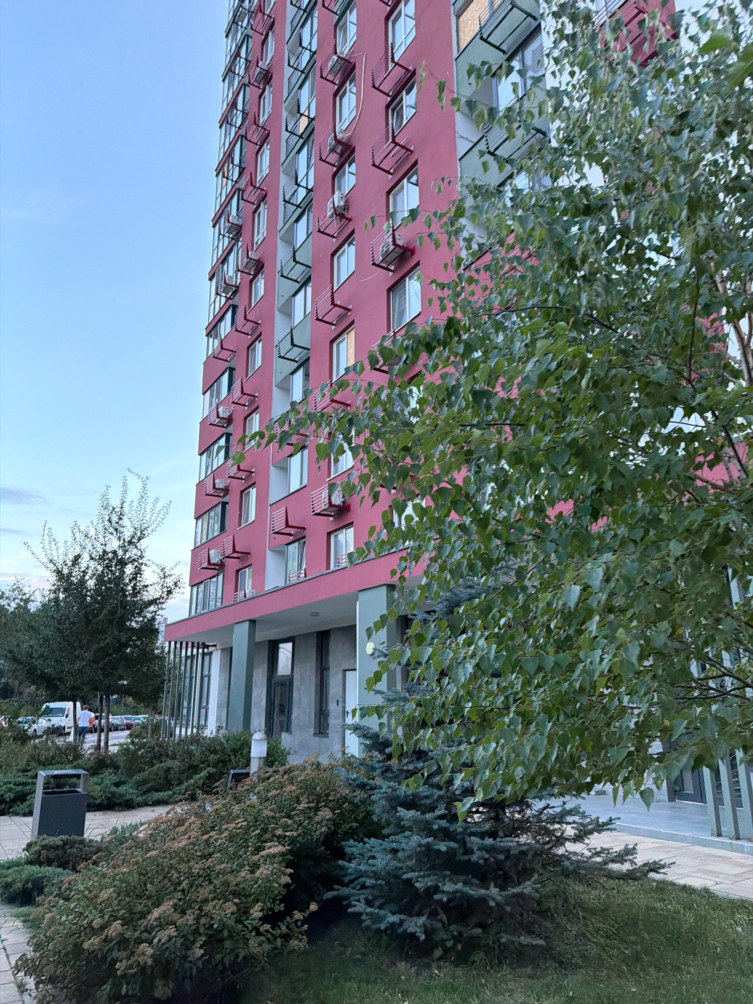 An outdoor, eye-level shot captures a tall, modern apartment building with a striking pink facade under a light blue, overcast sky. The building features rows of identical windows, many equipped with small, red metal air conditioning units mounted on brackets. A lush, green tree with dense foliage partially obscures the building from the right side of the frame. In the foreground, a variety of green shrubs and a small blue spruce tree create a landscaped area with a paved walkway. A dark gray trash receptacle is visible on the left, near the walkway. In the background, several cars and a white van are parked on a street lined with more trees, suggesting an urban residential setting. The overall scene depicts a typical city streetscape in Kyiv, Ukraine.