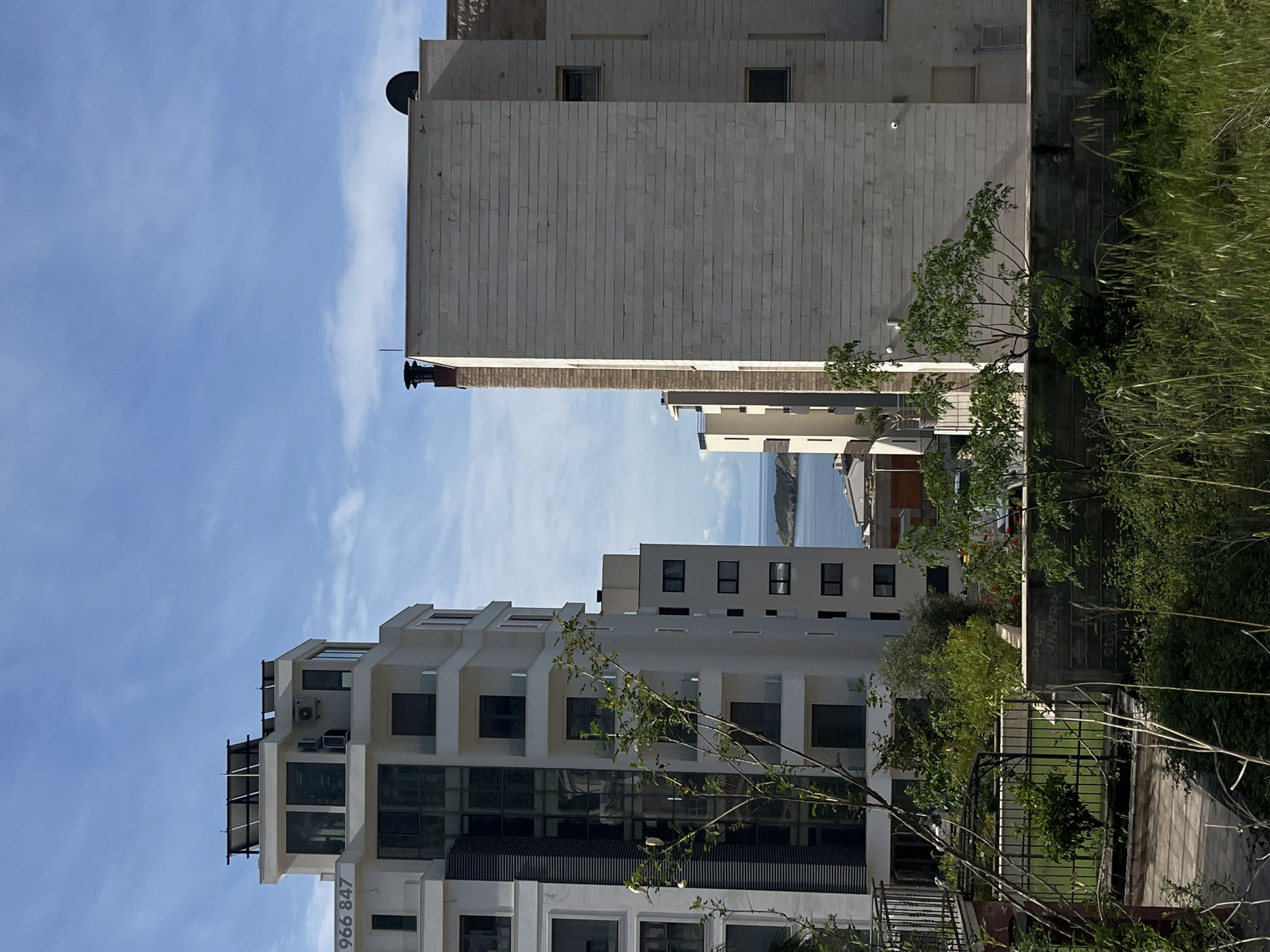 The image depicts a view of buildings and landscape in Boreti, Montenegro. In the foreground, a multi-story white building with balconies and glass windows is visible. To the right and slightly behind this building, a taller, blocky building with a light-colored brick facade stands. Between the buildings, a glimpse of the coastline and ocean can be seen in the distance. Lush green vegetation, including trees and grasses, grows on the right side of the frame, partially obscuring a pathway with a metal fence. The sky is a light blue with wispy clouds. A phone number, 966 847, is visible on the side of the lower building.
