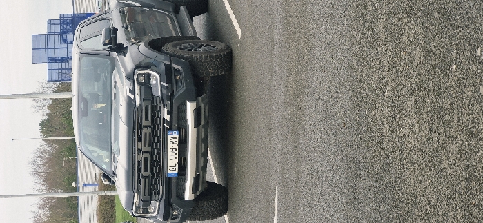 A gray Ford pickup truck, viewed from the front and side, is parked on asphalt. The truck has a large "FORD" grille, black wheels, and a license plate that reads "GL-506-RV." A blue metal structure appears in the background, along with some vegetation and a light-colored sky. The image is oriented horizontally.