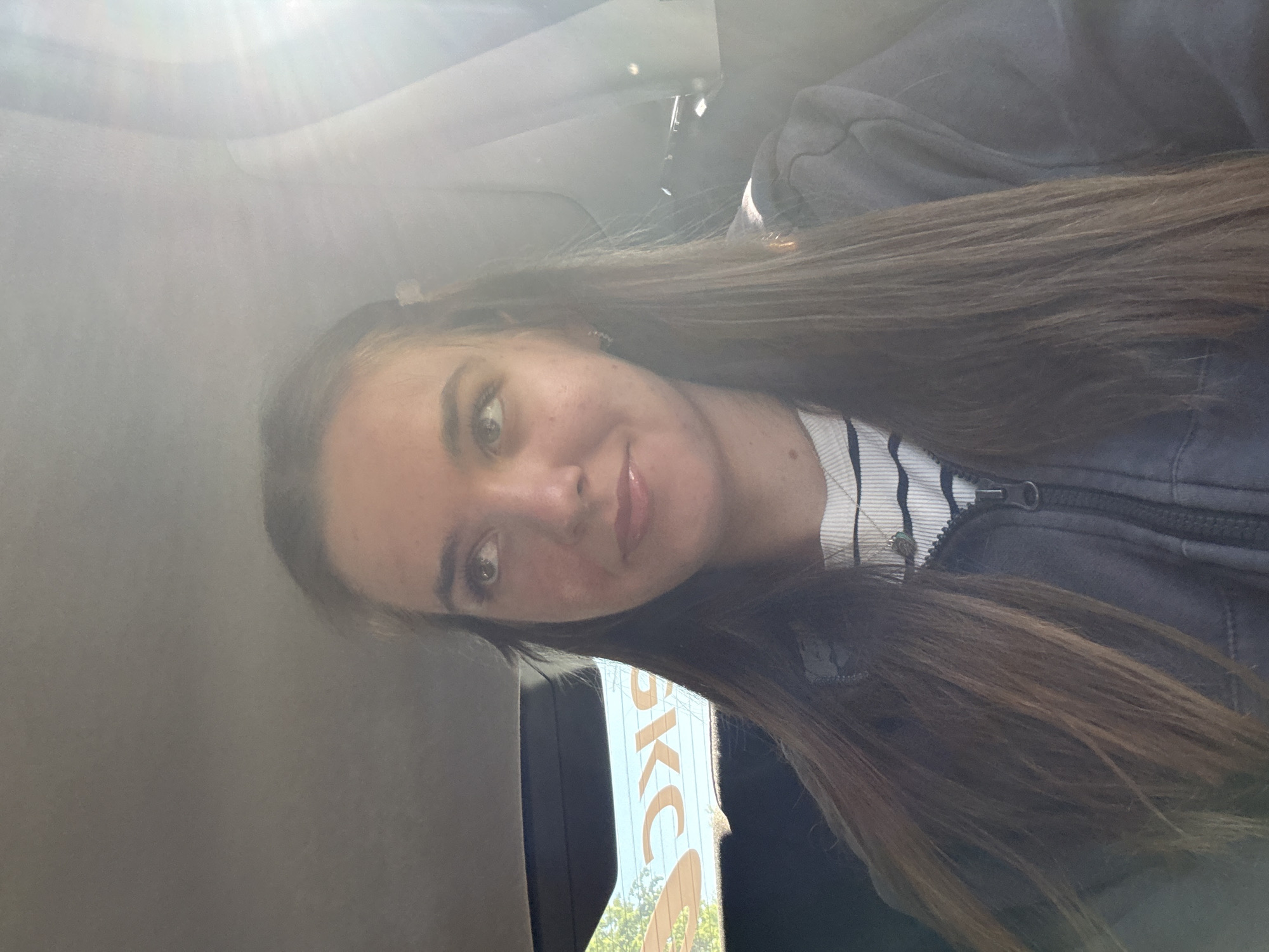 A selfie of a fair-skinned young woman with long brown hair. She is wearing a striped shirt under a dark zippered jacket. The photo appears to be taken inside a vehicle with a reflective interior. Outside the window, a sign with the letters "SKC" is partially visible, and this sign is associated with Perm, Russia. Sunlight is streaming into the frame, creating an overexposed effect in the top portion of the image.
