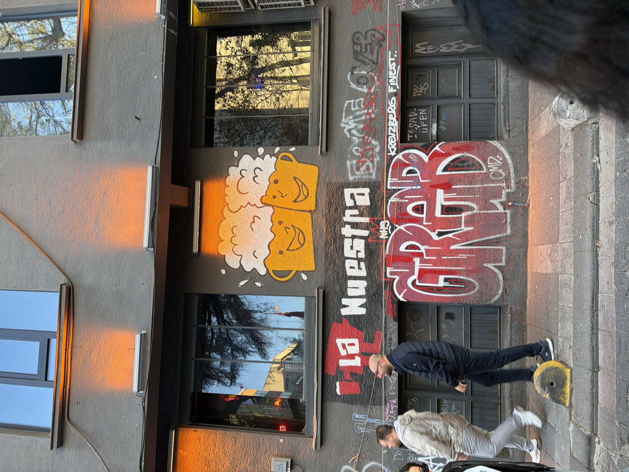 The image depicts the exterior of a building with graffiti in what seems to be an urban setting. There are two visible windows and a door covered with graffiti. The graffiti includes a drawing of two mugs of beer with smiling faces, the words "La Nuestra" in red and white, and the word "GRAB" in red. There are also other tags and markings. Two men are visible walking in front of the building. The environment appears to be a street with a sidewalk. The location cannot be accurately determined as Kadıköy, Turkey based solely on the provided image.
