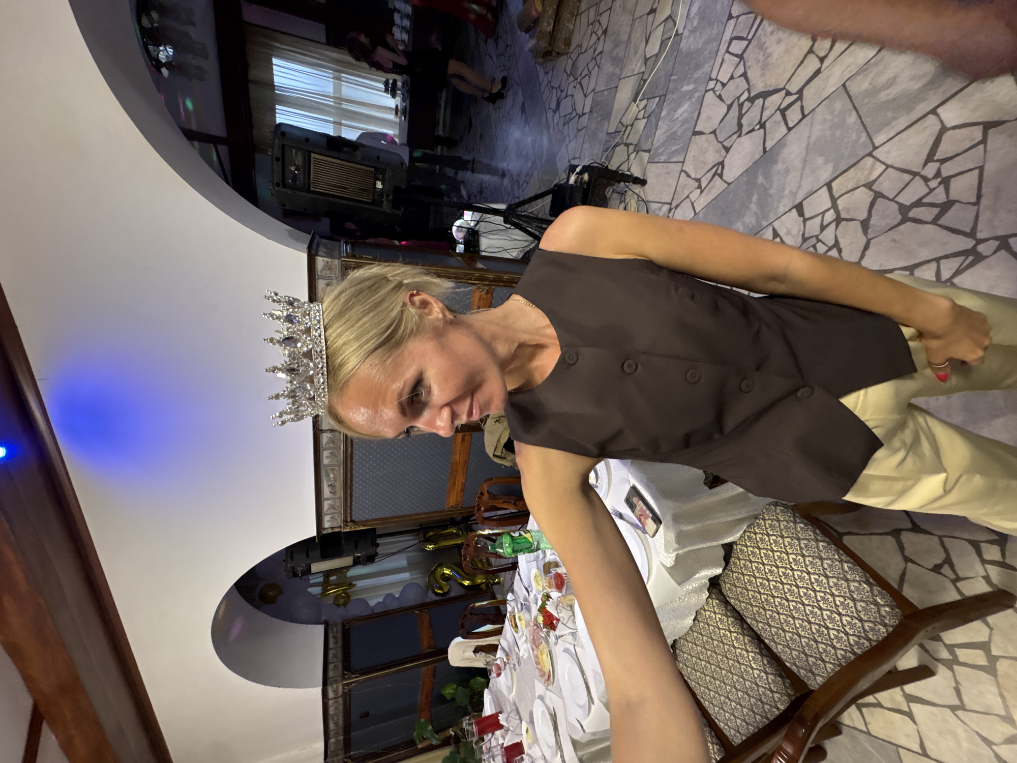 A woman wearing a silver crown and a brown sleeveless vest over a light-colored shirt stands in what appears to be a banquet hall in Zavod Mikhailovskii, Russia. She is looking to her left, with her arm extended forward. A table with white tablecloths is set for a meal, with plates, food, and drinks visible. Several chairs with patterned upholstery are around the table. In the background, gold balloons spelling out "21" are hanging, suggesting a birthday celebration. Other people are visible in the background, including a woman in a black dress. The room has arched doorways and is decorated with framed artwork. The floor is tiled in a mosaic pattern. A blue light emanates from the ceiling.