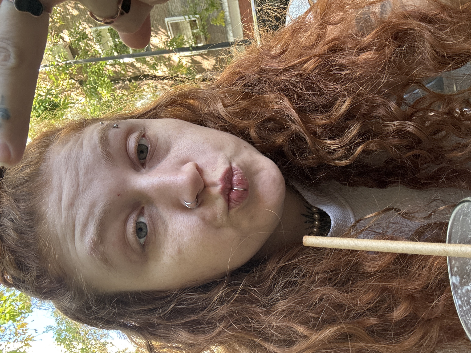 A young person with long, voluminous, curly reddish-brown hair is depicted in a close-up, eye-level selfie, angled slightly from above. The person has fair skin, light-colored eyes (appearing greenish-grey), and several facial piercings: two silver studs on their right eyebrow, a silver hoop in their left nostril, and a silver hoop on their lower lip. Their lips are slightly pursed, forming a pout, and they look directly at the camera.
A hand is visible in the upper left corner of the frame, holding what appears to be the camera, with rings on the fingers and a subtle tattoo on the side of the hand/wrist area. The person is wearing a white ribbed top and a beaded necklace. To the right, a light-colored straw is visible, extending from what seems to be a glass containing a drink.
The background suggests an outdoor setting during daytime, with bright natural light illuminating the scene. Green foliage from trees or bushes is prominent, along with glimpses of a building structure, indicating a garden, yard, or patio in Buenos Aires, Argentina. The weather appears sunny. There is no visible text in the image.