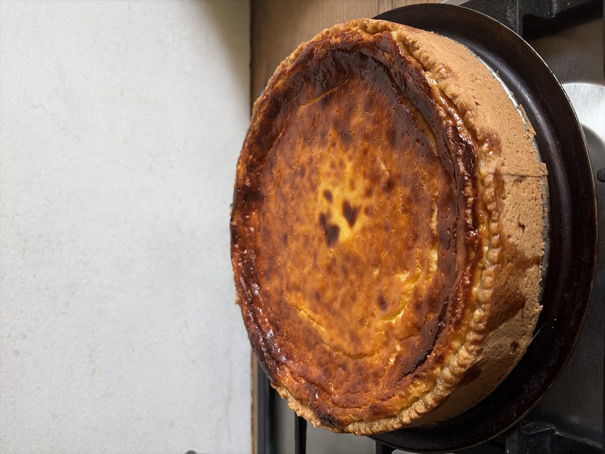 A close-up, high-angle shot shows a golden-brown, baked cheesecake on a dark circular pan. The cheesecake has a slightly puffed, caramelized top with darker browned spots and a golden, flaky crust. The cheesecake is positioned on a black stovetop with visible metal grates. In the background, a mottled white tiled backsplash is visible, and a wooden surface is to the right. The scene is indoors, likely a kitchen, and no people are present. The location is the city of Oxentea, Moldova.