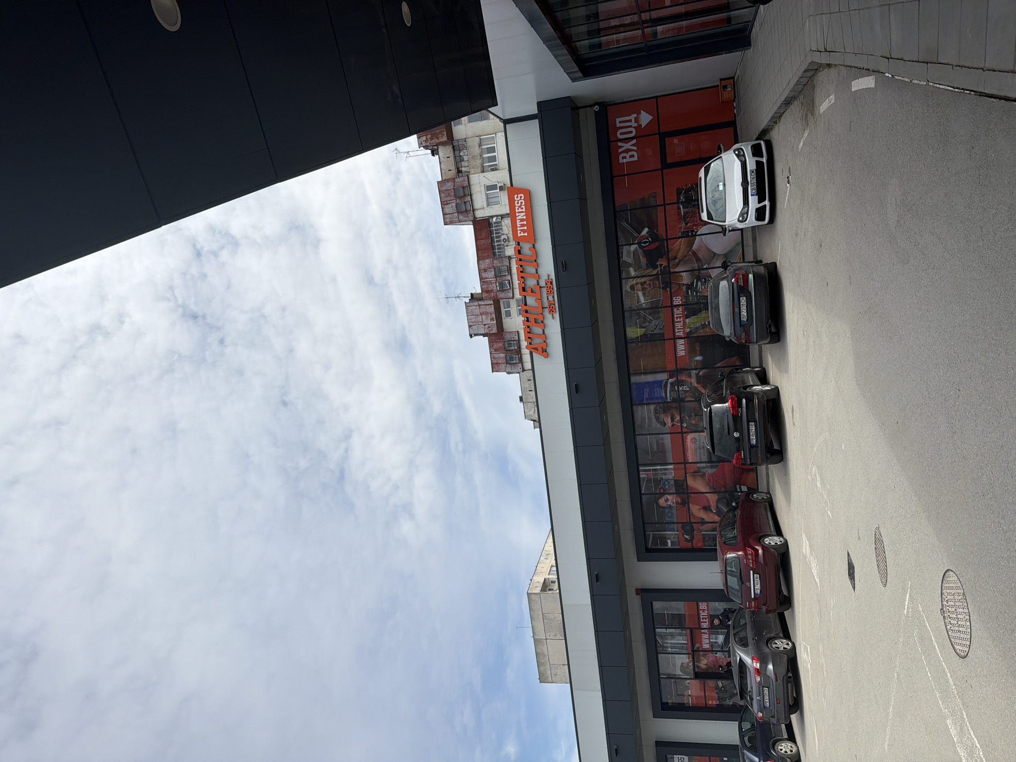 The image depicts the exterior of Athletic Fitness in Plovdiv, Bulgaria. The building features a modern facade with large windows displaying fitness-related imagery. A row of parked cars lines the parking area in front of the building. The sky above is partly cloudy. An older, multi-story building is visible beyond the fitness center. A large black awning is also shown.