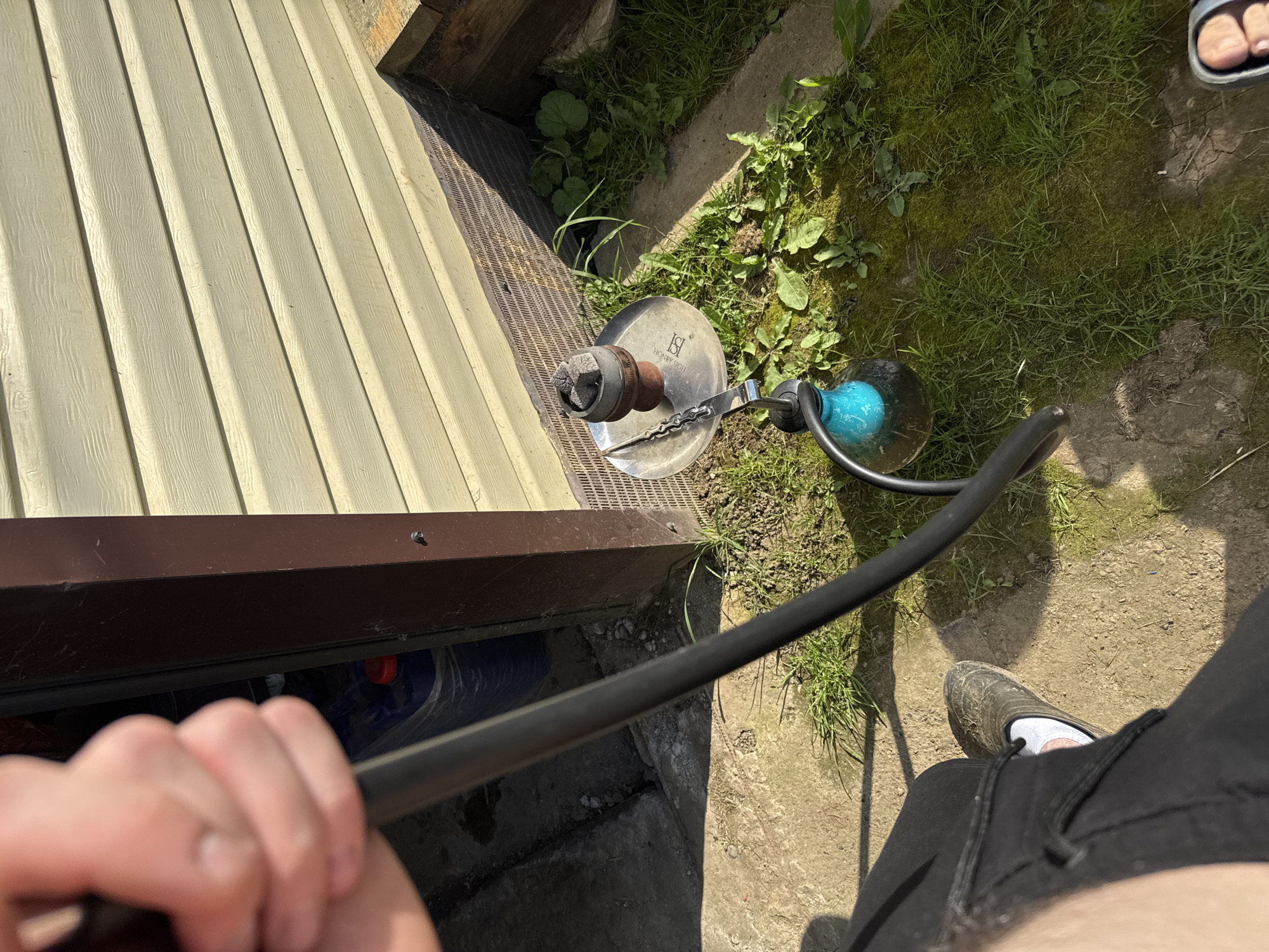 A person's hand holds a black hose, seemingly connected to a hookah. The hookah itself, with a blue glass base, rests on a patch of green grass next to a concrete surface. Above the grass, a light-colored wooden-paneled wall is visible, with a dark brown vertical beam running down the left side. The ground is a mix of dirt, grass, and concrete. The scene appears to be outdoors, possibly in a yard or garden area. There are no other people or animals visible in the frame. The lighting suggests it is daytime.