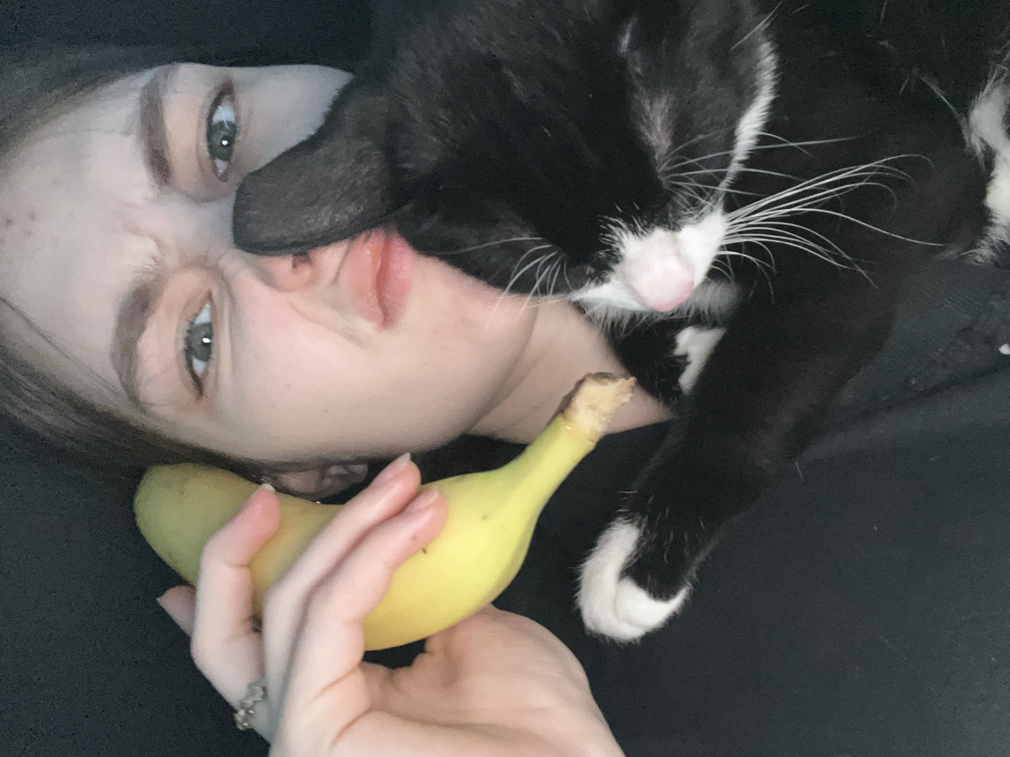 A woman is lying on a black surface, holding a banana in her right hand. A black and white cat is lying on her face, its head near her mouth. The location is not discernible from the image, and there is no indication of the city of Saint Petersburg, Russia.