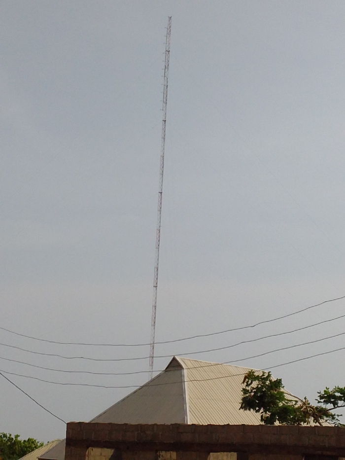 The image displays a tall, slender, reddish-brown lattice telecommunication tower positioned vertically against a light, uniform grey sky in Ajaokuta, Nigeria. The tower extends from the mid-frame to near the top edge. In the lower-midground, five dark, parallel utility lines span horizontally across the scene. Below these lines, the upper section of a building is visible, featuring a light-beige corrugated metal hip roof with sharp angles. Beneath the roof, a section of an unplastered reddish-brown brick wall is apparent. Green foliage is present to the right of the building's roof and faintly in the lower-left corner. No individuals or specific activities are depicted.