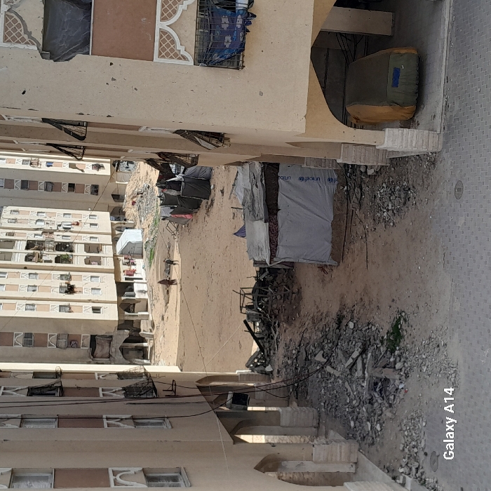 A dilapidated courtyard sits between multi-story residential buildings. Tents and makeshift shelters, some branded with UNICEF, are scattered across the sandy terrain alongside debris. Burnt remnants of structures lie in the foreground, suggesting recent damage. The scene conveys a sense of displacement and devastation in a densely populated urban area.