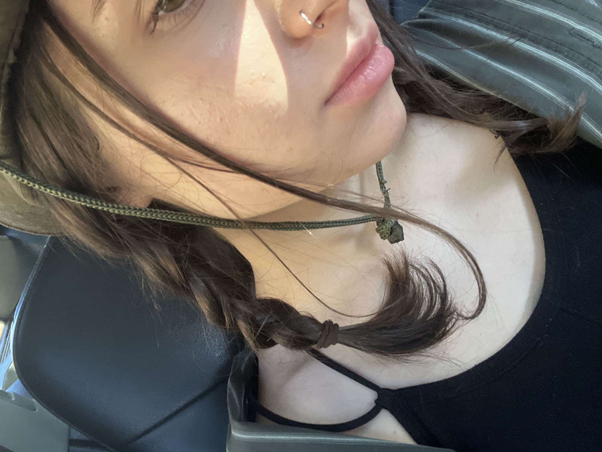 In a close-up, over-the-shoulder shot taken in the city of Tatariv, Ukraine, a young woman with long, dark brown hair braided into a single plait, is visible. She is wearing a black tank top and a greenish-gray striped jacket, possibly a hat with a cord. A green cord necklace with a dark stone pendant rests on her chest. Her right eye is visible, a light green iris, and her nose has a small silver hoop piercing. Sunlight casts a warm glow across her face, highlighting her skin and lips. The background shows a dark, upholstered seat, suggestive of a car interior. The overall mood is casual and personal, with the focus on the woman's features and attire.