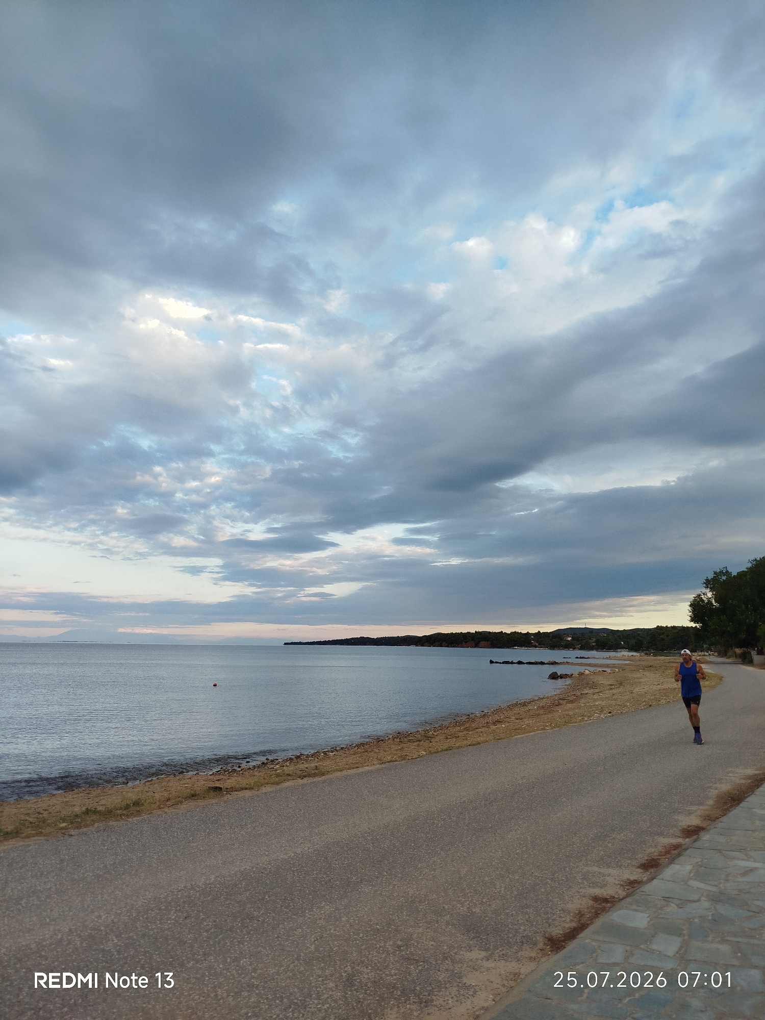 A person is jogging on a paved path next to a beach. The beach has sand and rocks, and the sea is calm. The sky is cloudy. A red buoy is visible in the water. The date and time stamp indicates it is July 25, 2026, at 7:01 AM.