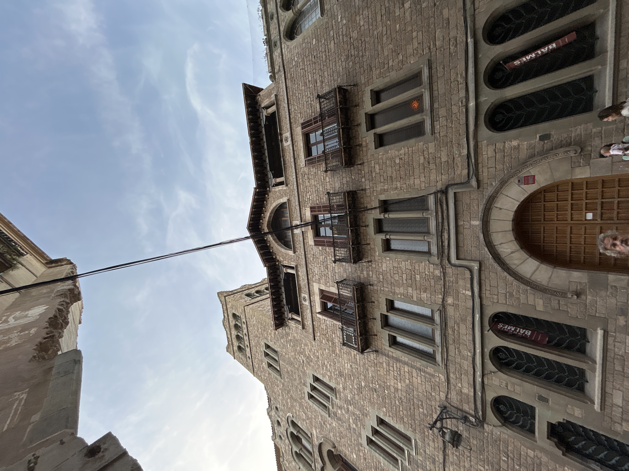 Captured from a low angle looking upwards, the image showcases the impressive façade of a multi-story building, likely in an urban European setting, consistent with Barcelona, Spain. The building, constructed from light-colored stone or brick, dominates the right side of the frame, featuring numerous windows of varying shapes. Some windows are rectangular, while others, especially on the lower levels, are distinctly arched and adorned with intricate dark metal grates. Several windows on the upper floors are fronted by small, ornate metal balconies or railings.
Towards the bottom right, a grand arched wooden doorway, possibly an entrance, is visible. Above a set of three arched windows, a red sign with white lettering clearly reads "LLIBRERIA BALMES," indicating a bookstore. The left side of the frame shows a partial view of another older, perhaps more weathered, building or wall.
The sky, visible in the upper portion, is bright blue with scattered wispy clouds, suggesting it is daytime. Two dark power lines or cables traverse the sky horizontally across the middle of the frame. A few indistinct figures of people can be seen near the main building's entrance on the far right, hinting at pedestrian activity. The overall impression is one of historical architecture and a lively city environment.