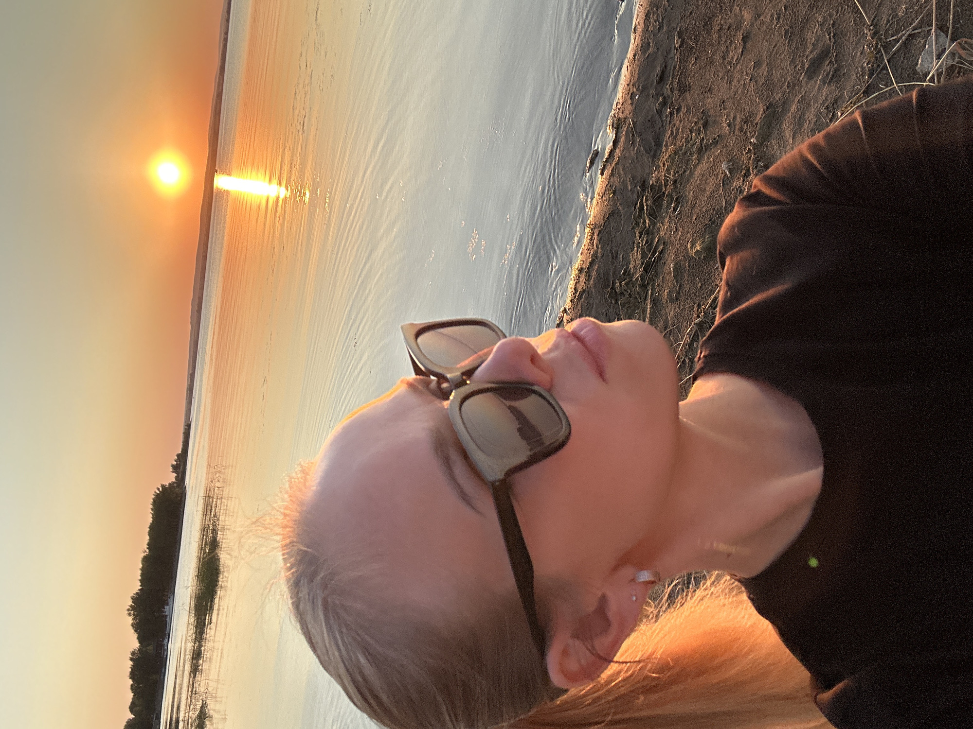 A young woman with blonde hair tied in a ponytail, wearing large, dark-rimmed sunglasses and a black t-shirt, is captured in a close-up, profile view. She is positioned on a sandy shore, looking out towards a large body of water. The sun is setting on the horizon, casting a warm, golden glow across the sky and reflecting brightly on the water's surface. The background features a treeline and a faint outline of distant land. The scene is likely located in Energetik, Russia, given the context of the provided image.