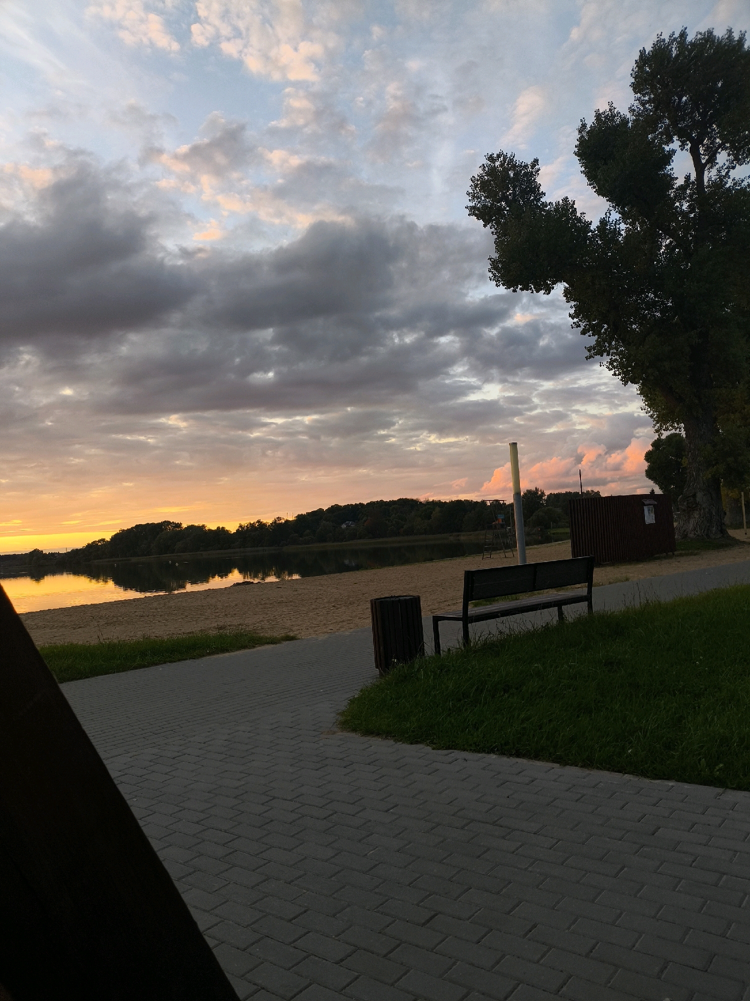 The image depicts a lakeside scene at dusk in the city of Myadzyel, Belarus. The sky is filled with dramatic clouds tinged with the orange and pink hues of sunset. A tranquil lake reflects the sky and the tree-lined shore. In the foreground, a paved walkway leads to a sandy beach. A dark-colored bench sits on the grass beside the walkway, adjacent to a trash receptacle. A tall, leafy tree stands prominently on the right side of the frame, its branches silhouetted against the sky. A wooden structure, possibly a shed or changing room, is visible behind the bench. The overall atmosphere is serene and peaceful.