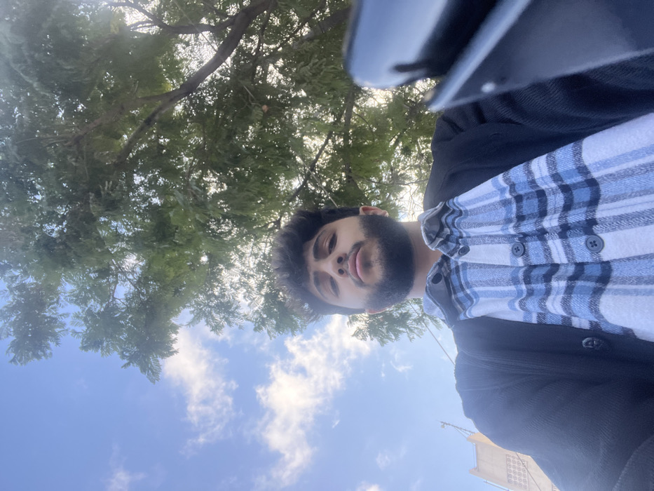A man with dark hair and a beard looks up at the camera from a low angle. He wears a black jacket over a blue and white plaid shirt. Behind him, a blue sky dotted with clouds peeks through the dense green leaves of a tree. Part of a black device is visible in the top right corner, suggesting the photo was taken with a phone. The bottom of a light-colored building is faintly visible.