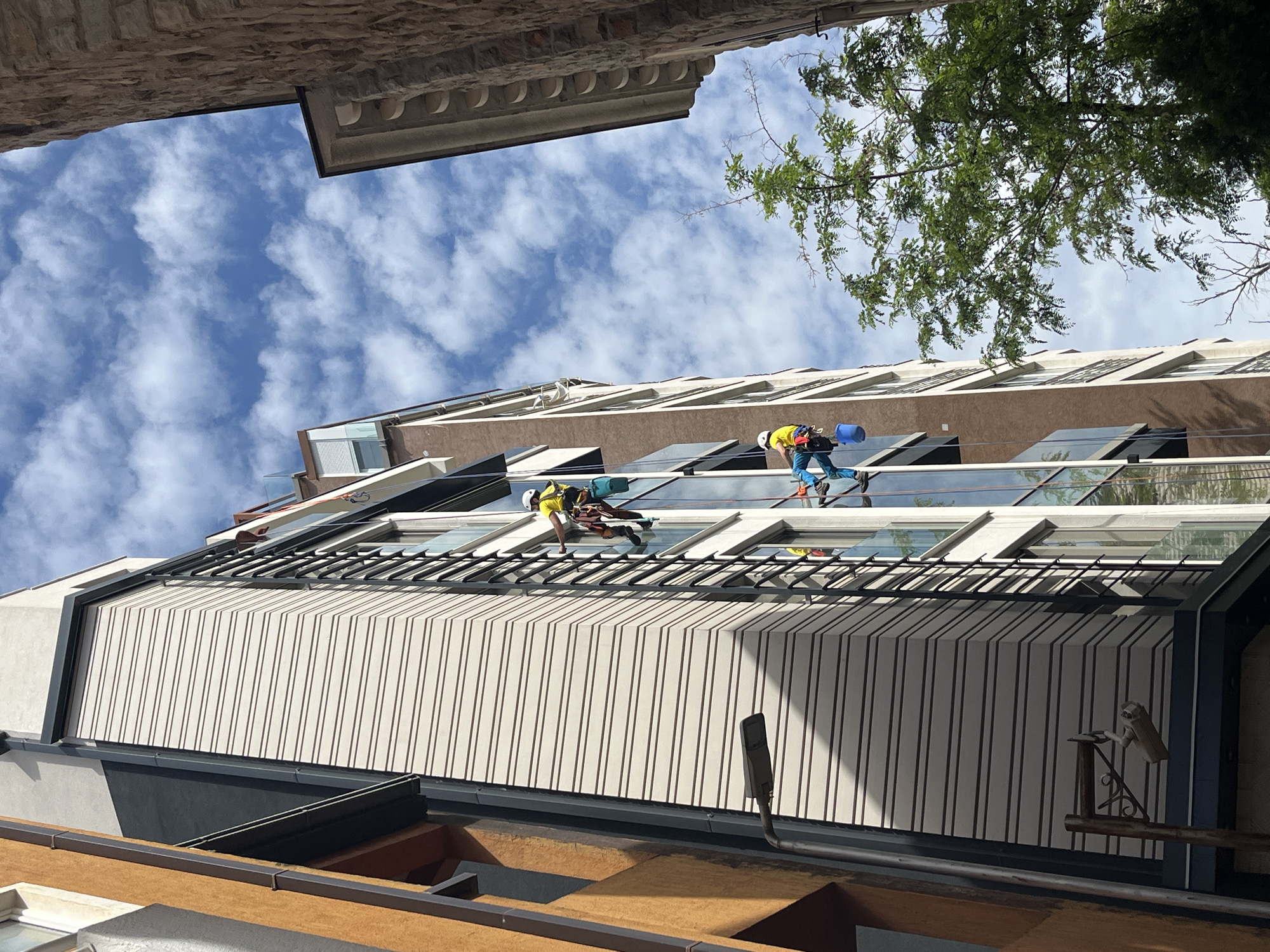 The image shows two window washers suspended on ropes cleaning windows on a modern multi-story building in Budva, Montenegro. The view is angled upwards from a lower building. The sky is partly cloudy. Building materials include beige and gray colored materials. A tree branch is visible in the upper right of the frame, partially obscuring the sky.