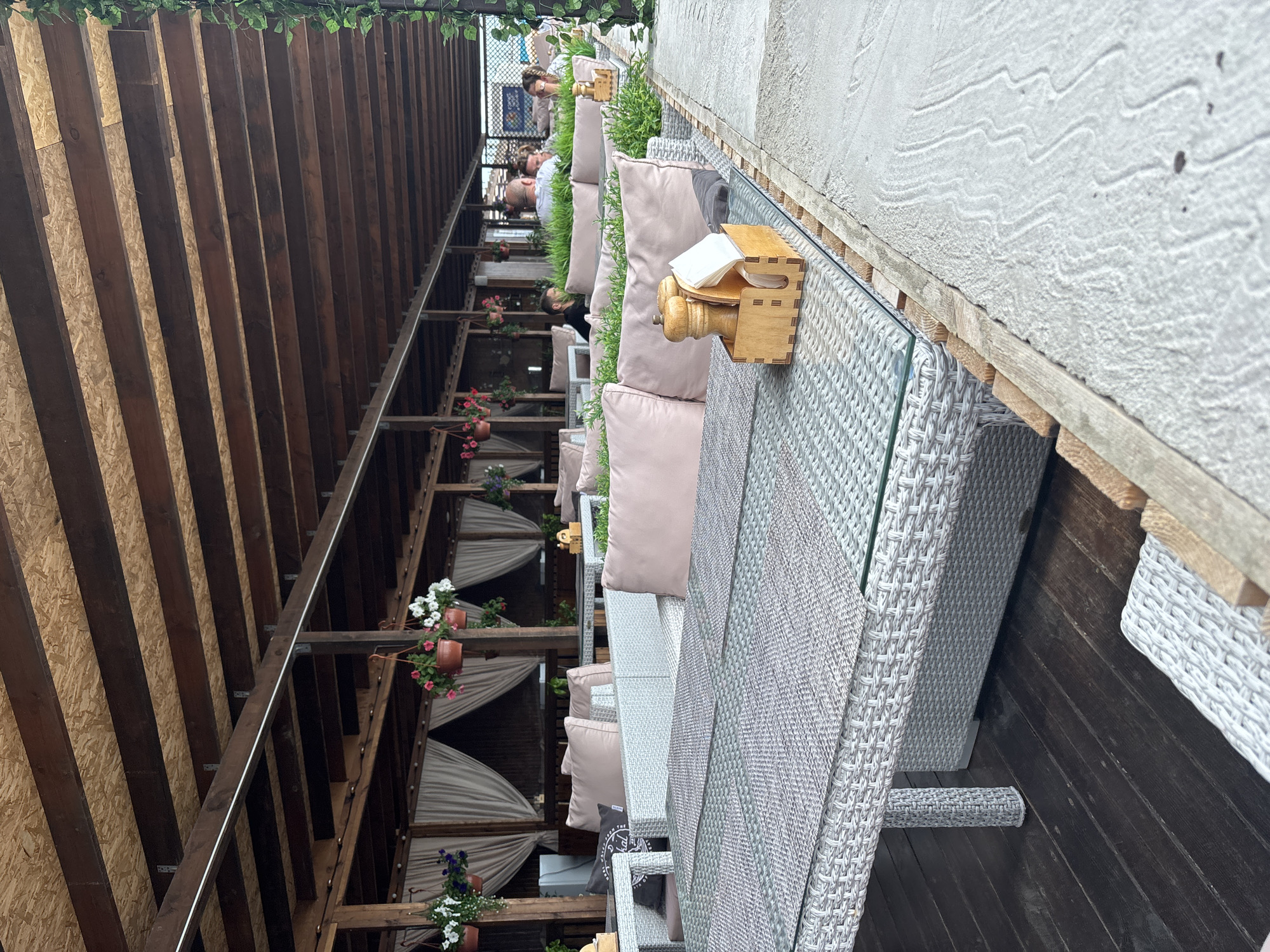 The image depicts an outdoor seating area, likely a restaurant or cafe, characterized by its covered wooden roof structure and rattan-style furniture. The furniture includes tables with glass tops and cushioned seating in a neutral pink hue. Hanging flower baskets are visible, adding a touch of greenery and color. In the background, several patrons are seated, engaged in conversation and dining. The flooring appears to be a combination of dark wood planks and a textured light-colored material. The scene is set during daylight hours, with soft lighting filtering through the roof. The overall ambiance suggests a relaxed and comfortable dining experience. The location is identified as a cafe in Sochi, Russia, implying a coastal or resort setting.
