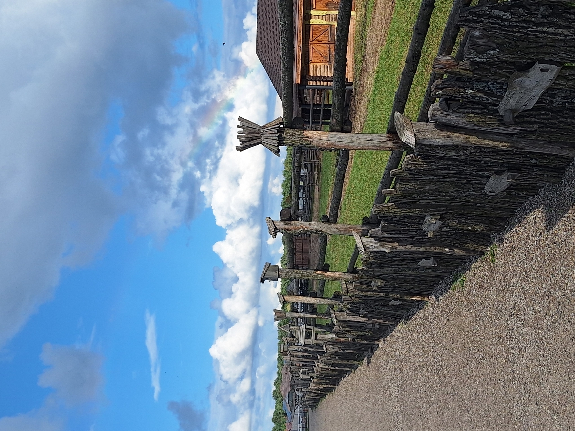 A rustic, wooden fence, made of rough-hewn logs and branches, runs diagonally from the lower left corner to the upper right of the image. The fence is old and weathered, with some sections appearing to be constructed from tree stumps and bark. Small, wooden birdhouses or feeders are attached to some of the fence posts. Beyond the fence, a grassy field stretches out, dotted with more wooden structures and fences. In the background, a cluster of buildings, likely farm buildings or rustic cabins, are visible, with a dark, shingled roof on one of them. The sky above is a bright blue, with fluffy white clouds and a faint rainbow arcing across the upper right portion of the frame. The ground in the foreground is a gravel path. The scene depicts a rural or historical setting, possibly an open-air museum or a farmstead, in the Malakhovetsky Selsoviet, Belarus.