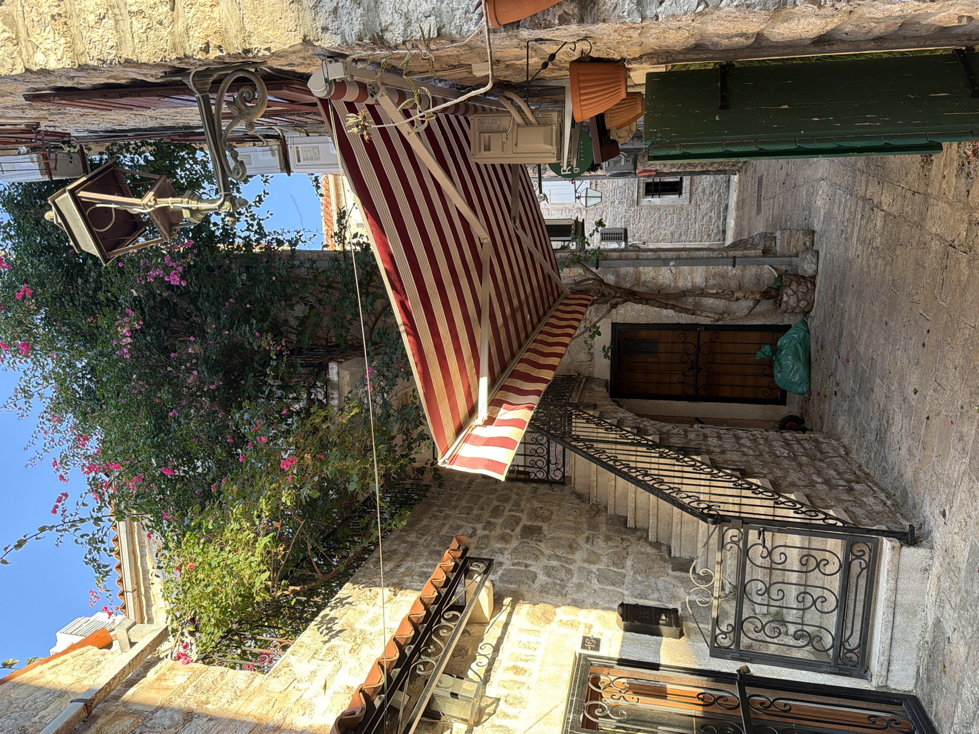 The image depicts a narrow cobblestone alley in the Old Town of Budva, Montenegro. The architecture is characterized by stone buildings with terracotta tiled roofs. In the foreground, a set of stone stairs with a wrought-iron railing leads upwards. To the left of the stairs, a metal gate with intricate scrollwork guards another entrance. Above the stairs, a red and white striped awning extends outwards from a building. A vintage-style lamp is mounted on the stone wall to the right of the awning. Lush green foliage with pink flowers cascades down from balconies and walls, adding a touch of color to the scene. A green wooden shutter is visible on the far right. The lighting suggests it is late afternoon, with strong shadows cast by the buildings and the awning. There are no people or animals visible in the frame.