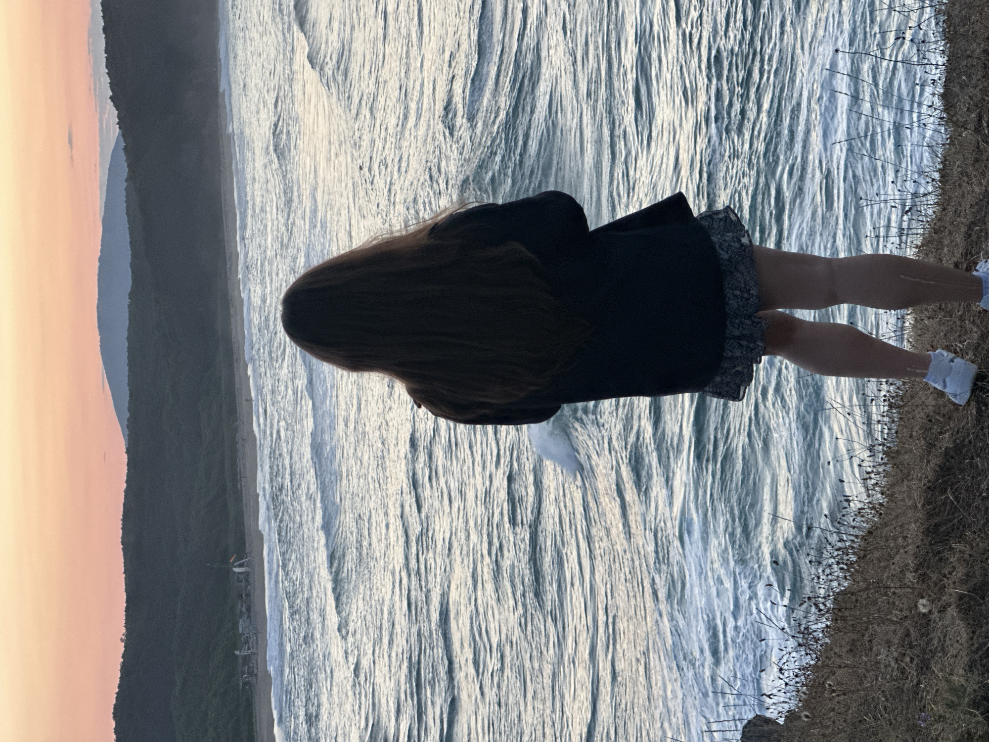 In this image, a person stands on a cliff overlooking a turbulent sea with crashing waves. The person, viewed from behind, has long, dark hair and is wearing a dark coat and a patterned skirt. Their legs are visible, and they are wearing white sneakers. The setting is a coastal landscape, with dry grass and plants on the cliff edge. In the background, a beach and a forested hill are visible, and the sky shows a gradient of orange and pink hues, suggesting either sunrise or sunset. The overall scene evokes a sense of contemplation and awe inspired by the natural beauty of the ocean and the rugged coastline. The image appears to be taken in Sinemorets, Bulgaria, given the visual characteristics of the coastline and vegetation.