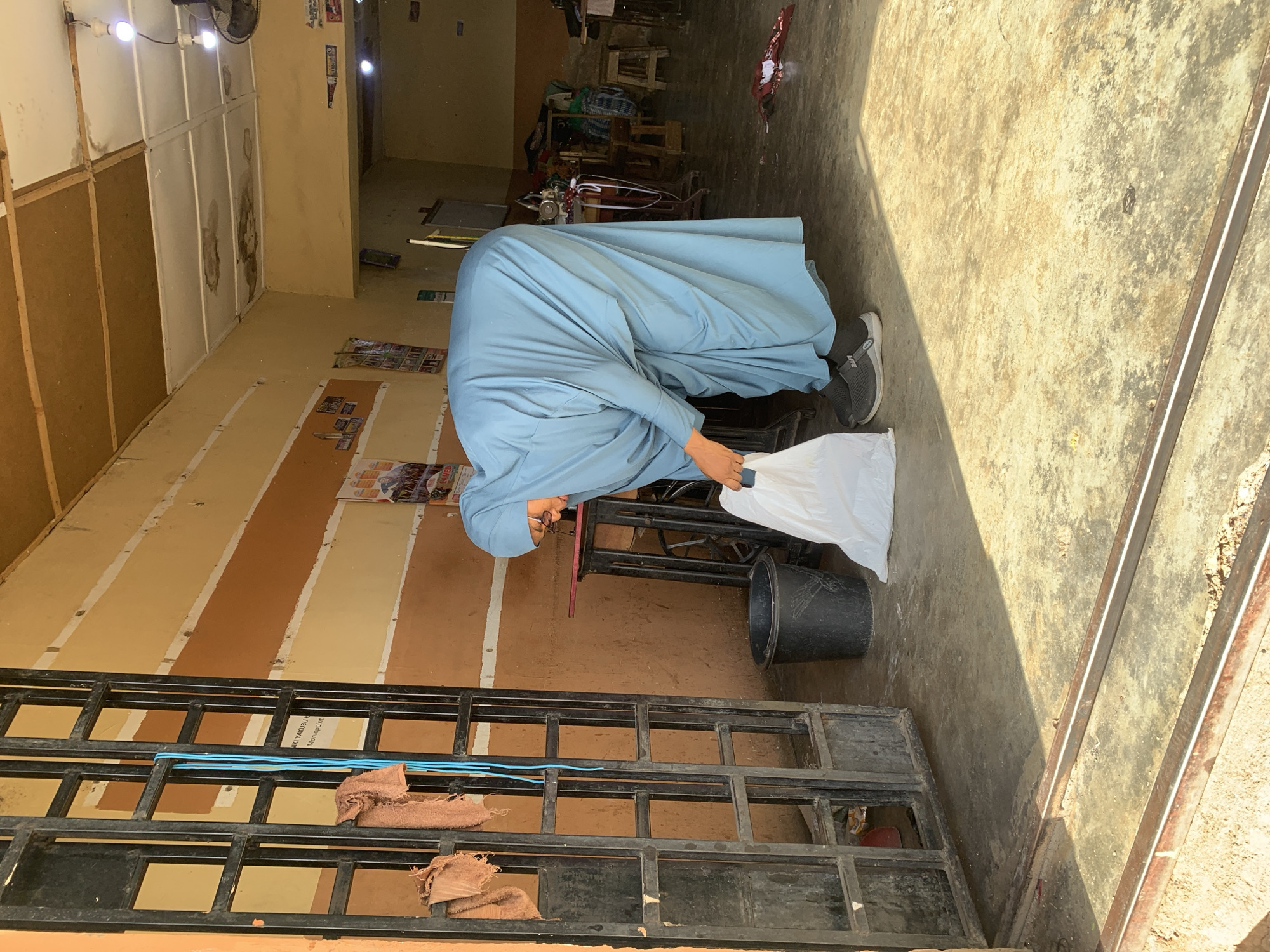 A man wearing a light blue robe and dark gray shoes is bending over while holding a white plastic bag in a dimly lit indoor space. The location appears to be a shop in Jos, Nigeria, based on signage visible on a promotional flyer attached to the wall. The interior features walls painted in horizontal stripes of tan and brown. A black metal gate lies on the floor in front of the man. Behind him, there's a sewing machine table with a black bucket nearby. In the background, are chairs and tables. The floor is concrete, partially lit by sunlight.