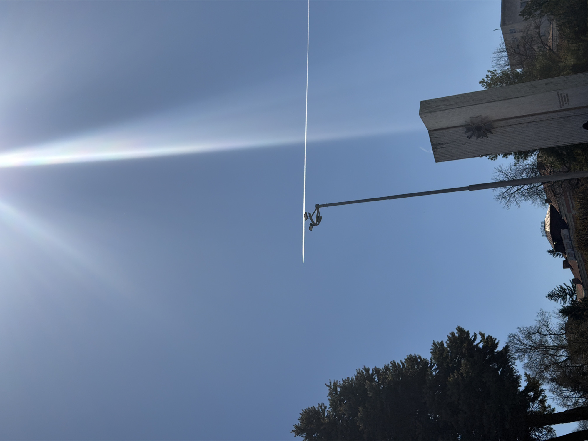 The image, taken from a low angle, shows a bright, blue sky with the sun shining from the upper left corner. A white contrail runs vertically through the center of the frame. In the midground, there is a street lamp post and a rectangular stone monument featuring a floral or rosette design. Trees are visible at the bottom and partially on the right. The location is likely the city of Pleven, Bulgaria.