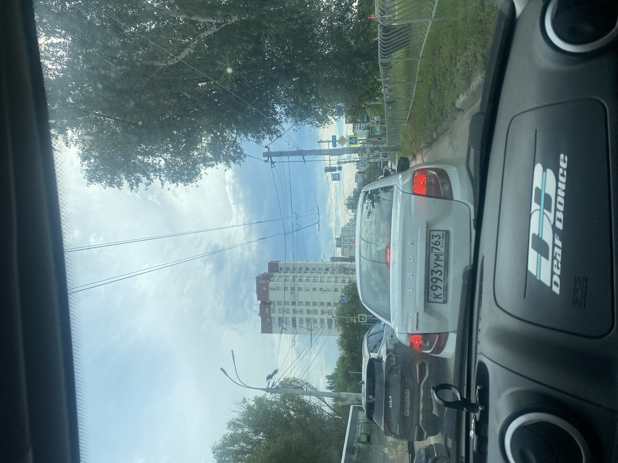 The image captures a view from inside a vehicle, looking out at a city street in Samara, Russia, during daylight. The scene is dominated by traffic, with several cars visible on the road. In the foreground, the dashboard of the vehicle is visible, including part of a steering wheel and a decorative sticker that reads "DEAF DANCE".
Further ahead, a white LADA Granta is the most prominent vehicle, displaying a Russian license plate K993YM 763. To its left, a dark-colored Kia Sportage is also in traffic, with its license plate starting with M109BH. A silver sedan is partially visible behind the Kia.
The background features tall, multi-story apartment buildings, characteristic of urban Russian architecture. Overhead, a network of power lines and trolleybus wires crisscross the sky, indicating the presence of public transportation. Trees line the right side of the street, with dense foliage suggesting a summer season. The sky is partly cloudy, with patches of blue visible. Traffic lights and road signs are present at intersections, implying a busy urban environment. The overall impression is one of a typical traffic jam in a Russian city.