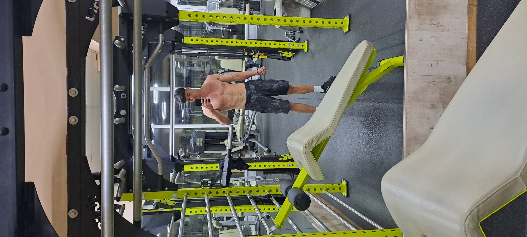 A shirtless man is standing sideways in a gym, holding up his phone with his left hand, presumably taking a photo of himself. He is wearing black patterned shorts, white socks, and black shoes. The gym is equipped with various weightlifting machines, including squat racks with yellow accents. A bench press with a light-colored seat is positioned at an angle. The gym has a rubber mat floor and mirrors along the walls. The location appears to be in a well-lit, commercial or private gym setting. While the image does not provide information about the city of Burgas, Bulgaria, the description remains focused on the physical elements and actions depicted in the scene.