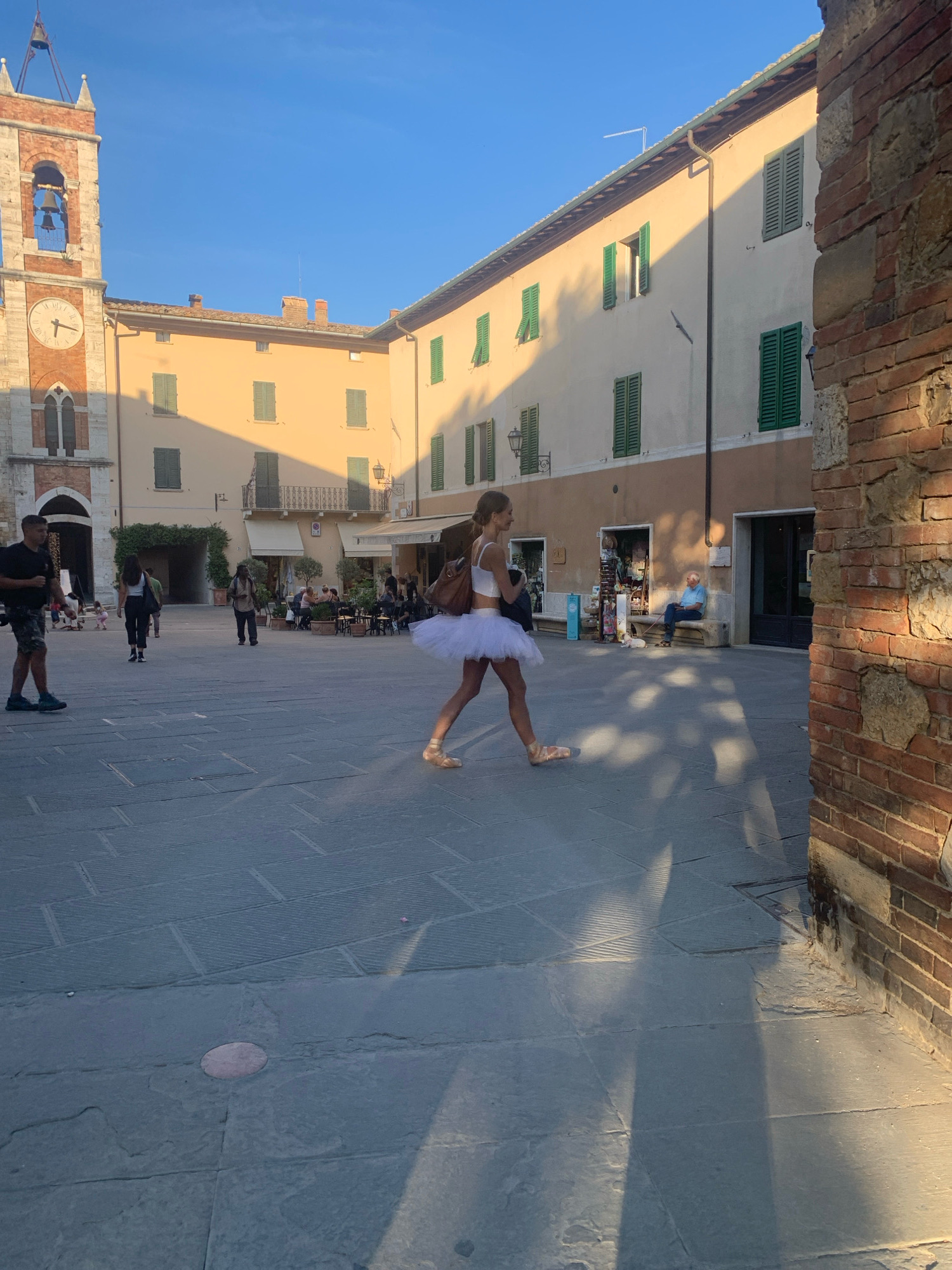 The image depicts a sunlit town square in San Quirico d'Orcia, Italy. In the foreground, a young woman wearing a white tutu and ballet slippers is captured mid-stride, her shadow stretching across the stone pavement. To the left, a clock tower with red brick accents and a bell is visible. Buildings with cream-colored facades and green shutters line the square, some featuring shops and outdoor seating for a cafe. Several other individuals are scattered throughout the square, walking or sitting. A man in a blue shirt sits on a bench near a doorway, accompanied by a small white dog. The overall scene suggests a warm, pleasant day in a historic Italian town.