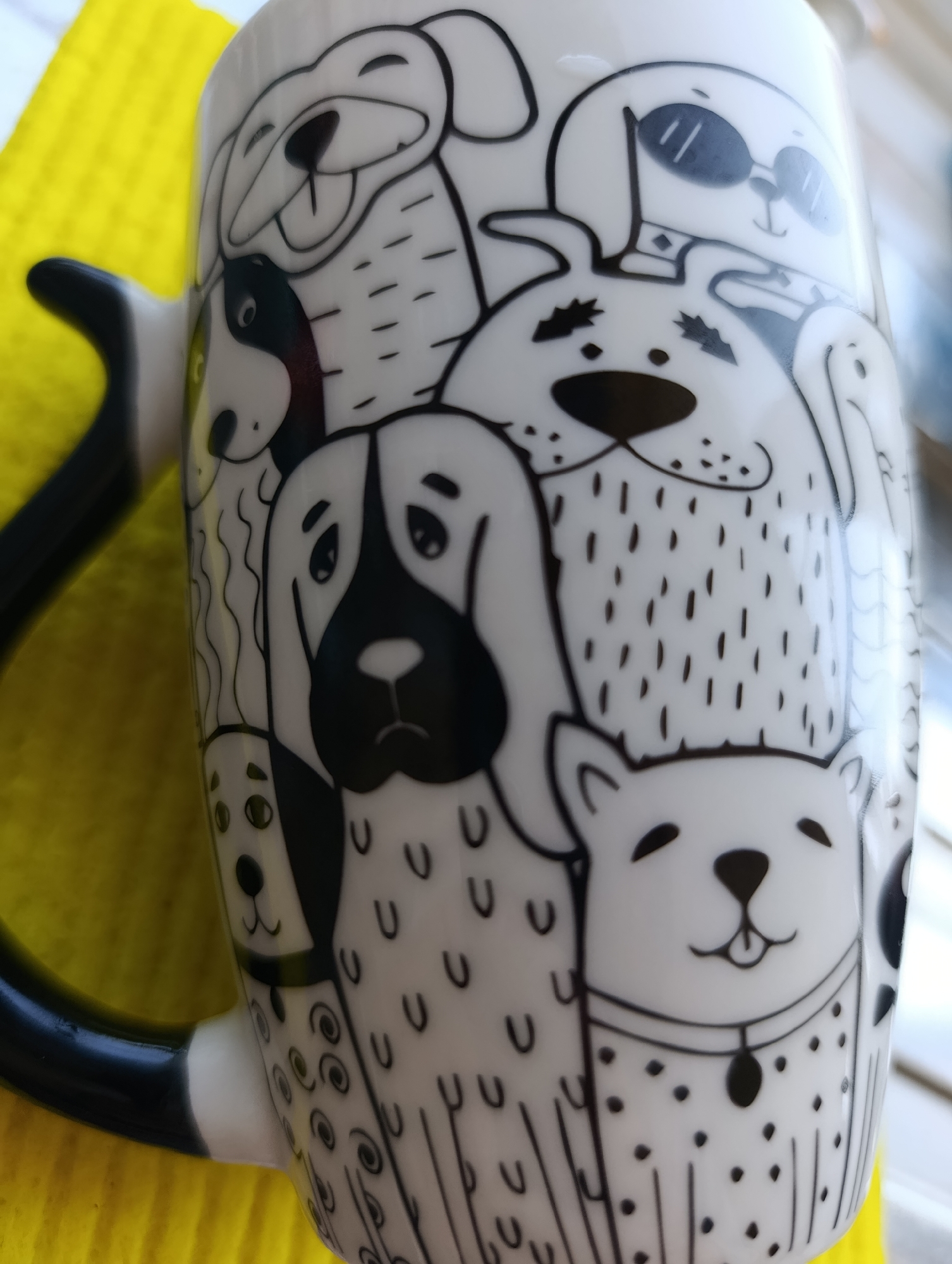 The image shows a white mug with black cartoon dog illustrations on a yellow surface. The mug handle is black with white ends. The illustrations depict various dog breeds and styles. The scene's location in Plovdiv, Bulgaria is not discernible from the image.