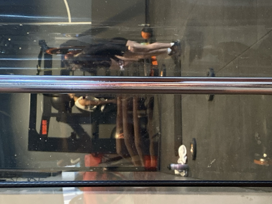 The image depicts an interior view of a Basic-Fit gym located in Hœnheim, France, captured through a reflective glass surface. A vertical, chrome-plated metal bar occupies the central foreground, partially obstructing the view.
Within the gym, two individuals are visible, likely reflected in the glass. On the left side of the reflection, a person wearing a white sleeveless top is partially seen from behind, standing near a black gym rack which bears the "BASIC-FIT" logo in red lettering. Below this, several horizontal, reddish-brown upholstered benches or steps are stacked on the dark floor.
On the right side of the reflection, another individual is standing, clad in a dark long-sleeved top and dark shorts, exposing their legs. They wear athletic shoes with white socks and are positioned facing left. On the dark gym floor in the foreground, near the left edge, a light purple water bottle rests beside a white towel and a dark weight plate. Another dark weight plate is visible further right on the floor, and a black spherical object, possibly a medicine ball, is on the floor near the right-side individual's feet. The walls are dark gray. The scene appears well-lit by interior lighting.