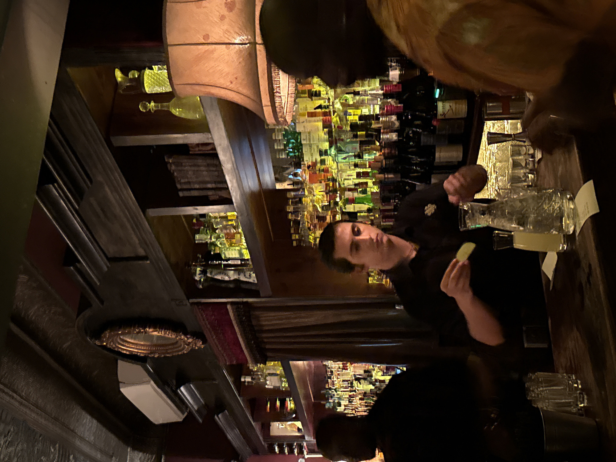 A dimly lit bar scene. A bartender, dressed in black, is holding a lime wedge. Multiple rows of liquor bottles line the back wall behind him. In front of him are cocktail glasses and a jigger, and a customer stands opposite him with a yellow patterned shirt. The location is likely a bar in Cape Town, South Africa.
