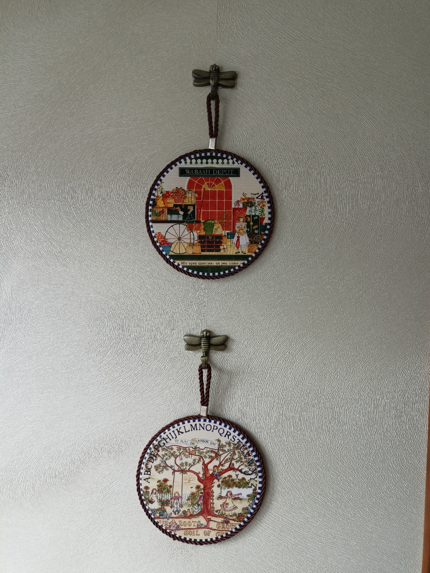 Two round, decorative wall hangings are displayed vertically on a textured, light gray wall. Each hanging is suspended from a metal dragonfly-shaped hook by a dark braided cord.
The upper hanging features a scene with the text "WABASH DEPOT" at the top. The artwork depicts a red arched doorway, a shelf with various items including baskets of fruit and gifts, and a woman standing with a doll. A spoked wheel is also visible. Below the image, the text reads, "His eyes upon you as you come."
The lower hanging is also circular and has text around its perimeter, including "A, B, C, D, E, F, G, H, I, J, K, L, M, N, O, P, Q, R, S." In the center of this hanging is a depiction of a tree with children playing in and around it, including on a swing. Text at the bottom reads, "OUR ROOTS GROW SOIL OF GOLD."
The wall has a consistent, ridged texture. The lighting appears to be from above and to the right, casting subtle shadows. There is a vertical wooden trim piece visible on the far right edge of the frame. The scene is static and shows no signs of activity. The location is unknown.