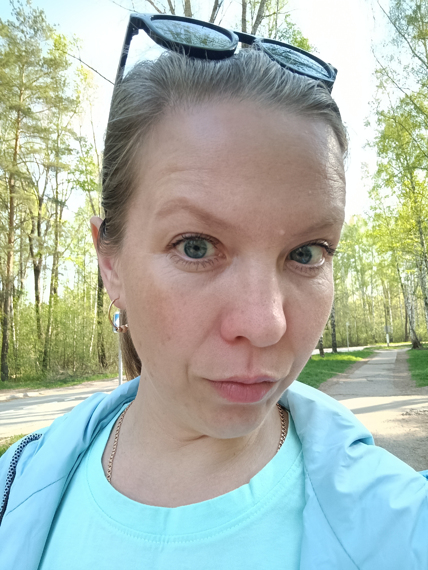A close-up selfie of a fair-skinned woman with blue eyes and sunglasses perched on her head. She is wearing a light blue t-shirt and jacket, along with a gold necklace and hoop earrings. The background shows a park setting with trees lining a paved path. Based on the location in Chaikovsky, Russia, it is likely a park or recreational area within the city.