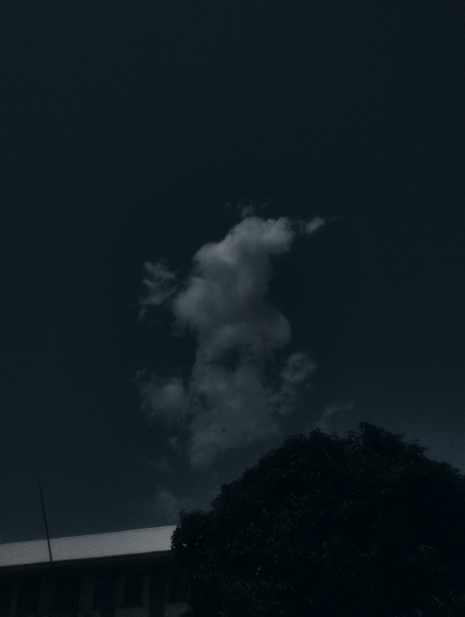 The image depicts a dark, moody sky with a solitary, textured cloud formation near the center. Below the sky, the silhouette of a large tree dominates the lower right portion of the frame. To the left of the tree, the roofline of a building is visible, appearing as a light-colored horizontal band against the dark sky. A thin, dark vertical pole extends upwards from the building's roof. The overall atmosphere is somber and potentially indicative of an approaching storm or twilight. The scene is set in Jos, Nigeria, suggesting a tropical or subtropical environment. The lighting and color palette, dominated by dark blues and grays, create a sense of mystery and perhaps a touch of melancholy. No people or specific activities are discernible.