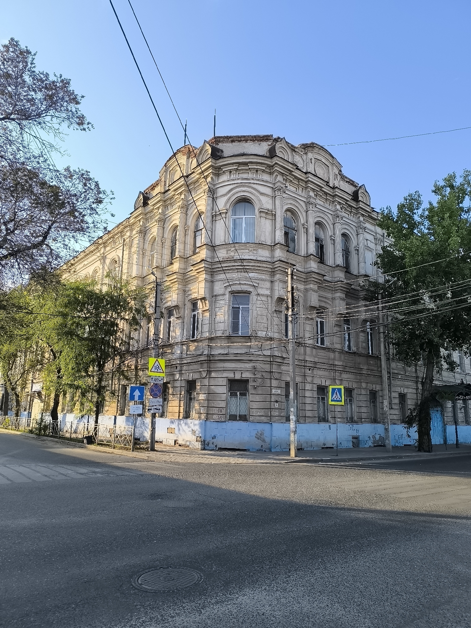 The image shows a street corner in Astrakhan, Russia, with a prominent, old, multi-story building. The building is an architectural style typical of the late 19th or early 20th century, with ornate detailing around the windows and cornices. The facade is weathered and shows signs of age, with peeling paint visible on the lower sections.
Several trees line the street, their branches casting shadows on the building and the road. Overhead, power lines crisscross the sky, typical of urban infrastructure.
On the sidewalk near the building, there are traffic signs, including a pedestrian crossing sign with a yellow triangle and a pedestrian symbol, and other directional signs. A blue-painted fence runs along the sidewalk in front of the building. The road in the foreground is asphalt and appears to be a street in the city, with a manhole cover visible.
The sky is clear and blue, indicating good weather. The lighting suggests it is either early morning or late afternoon, given the long shadows. There are no people or vehicles immediately visible in the frame, giving the scene a quiet, almost deserted feel. The overall impression is one of a historic urban environment showing signs of wear and tear but retaining its architectural character.