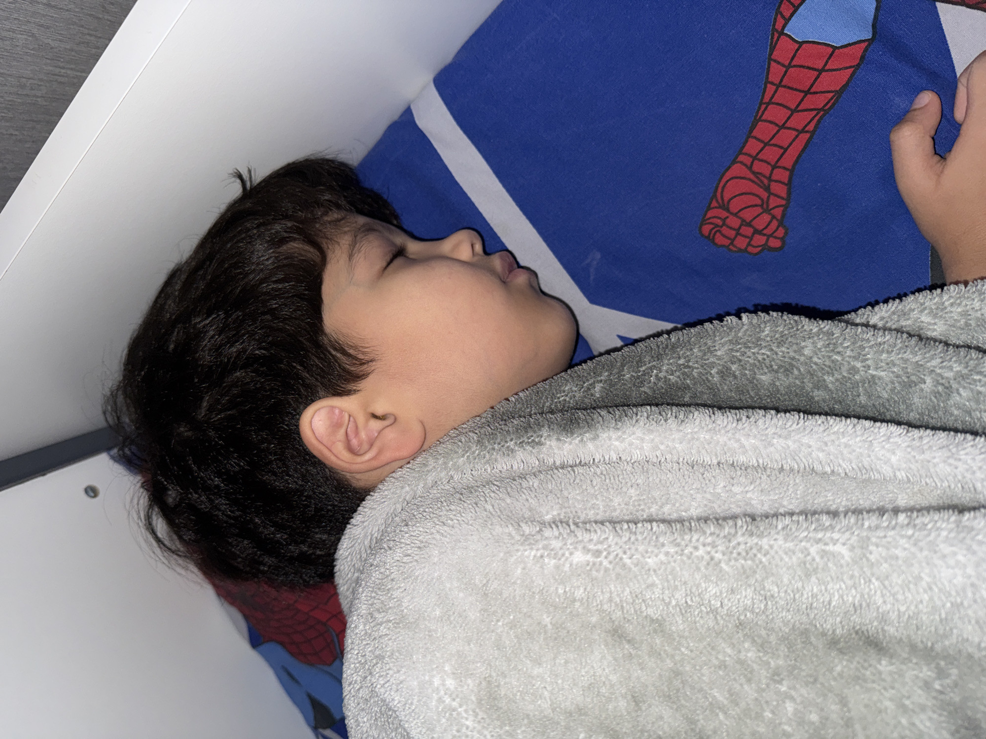 The image shows a young child sleeping, nestled on a bed. The child is lying on their side, covered by a soft, grey blanket. Their eyes are closed, and they appear to be in a deep sleep. The child has dark hair and is wearing a grey bathrobe.
The bedding features a vibrant blue pillowcase with a Spiderman comic character design, specifically the iconic red and blue suit with its web pattern. A portion of the Spiderman costume is visible, showing the textured red boot and part of the blue suit.
The background includes a white surface, likely a wall or the edge of a bed frame, with a subtle shadow. The lighting suggests it could be nighttime or a dimly lit room, contributing to the peaceful atmosphere of sleep. There are no discernible signs of text in the image. The overall scene evokes a sense of comfort and tranquility.