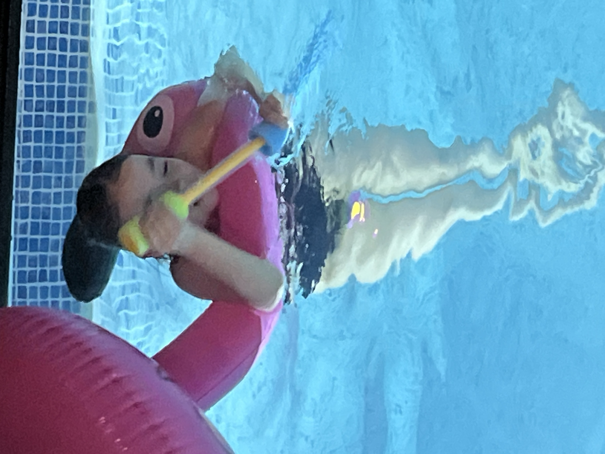 A child floats in a swimming pool on a pink flamingo float, holding a toy water gun. The pool has blue tilework along the side. Refraction of light through the water is visible. The location is unspecified; the city of Izhevsk, Russia, is not apparent.