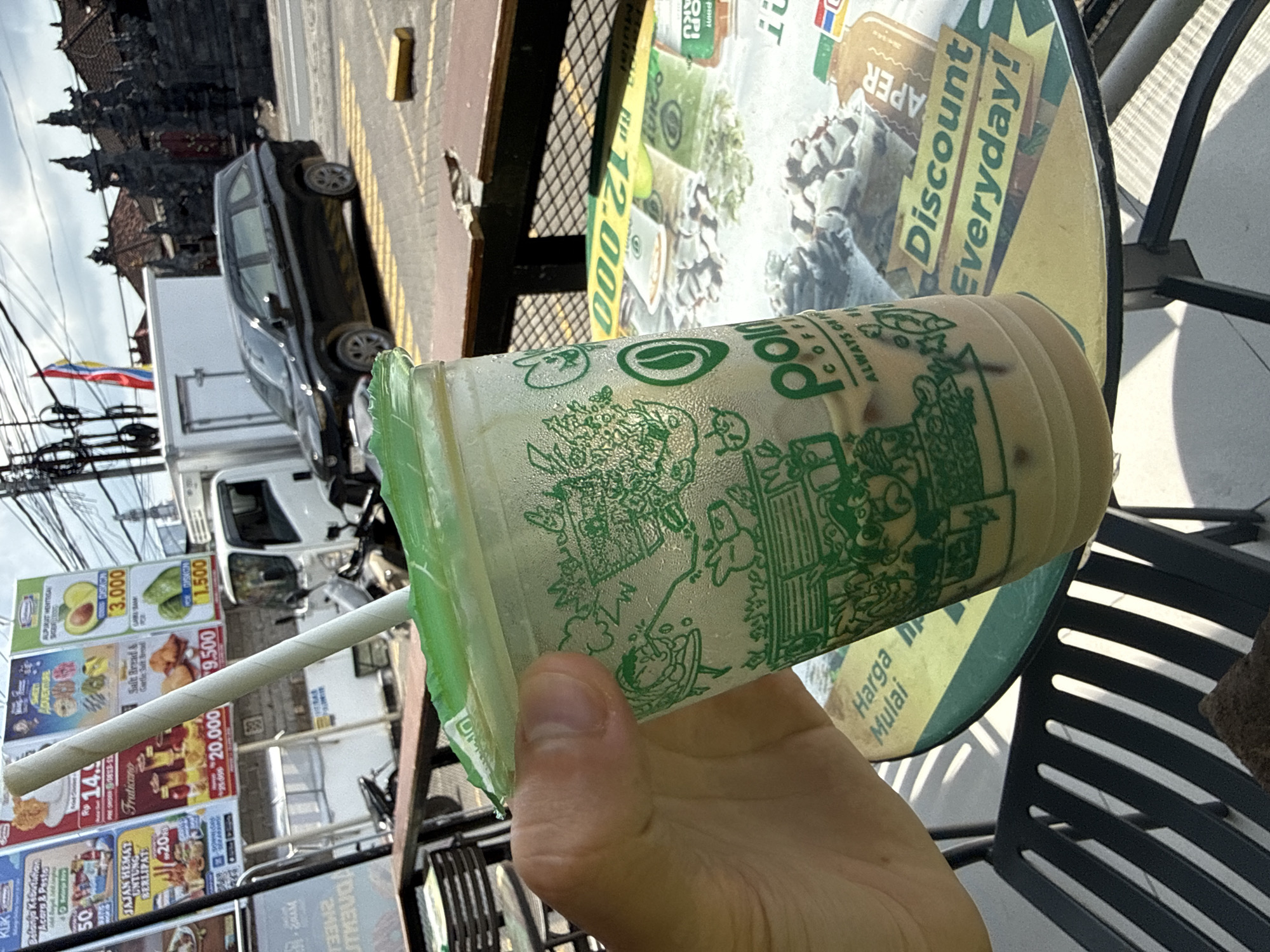 A person is holding a disposable Polos coffee cup filled with iced coffee on a table in Kutuh, Indonesia. The coffee cup has a green design on it and a straw. The table has a circular ad for the Aper discount. A black metal chair is visible. In the background, there are billboards advertising food items and prices, as well as a black car and a white delivery truck parked on the street. Power lines and buildings are also visible.