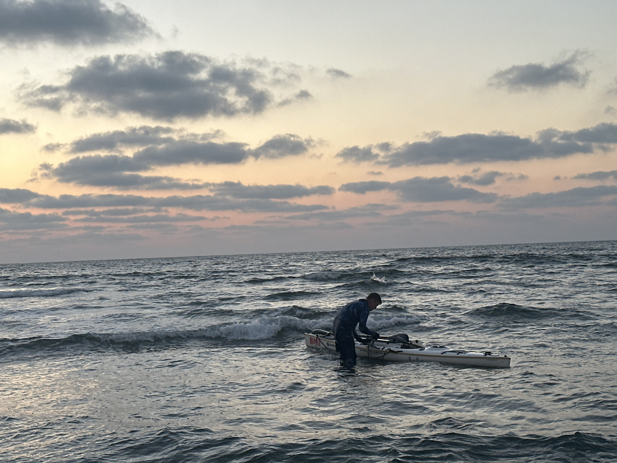 A person in dark clothing stands in the ocean near a white kayak, appearing to be preparing it or securing something. The water is choppy with small waves. The sky above is a pale gradient of grey and pink clouds, suggesting either dawn or dusk. The location is the coast of Haifa, Israel.