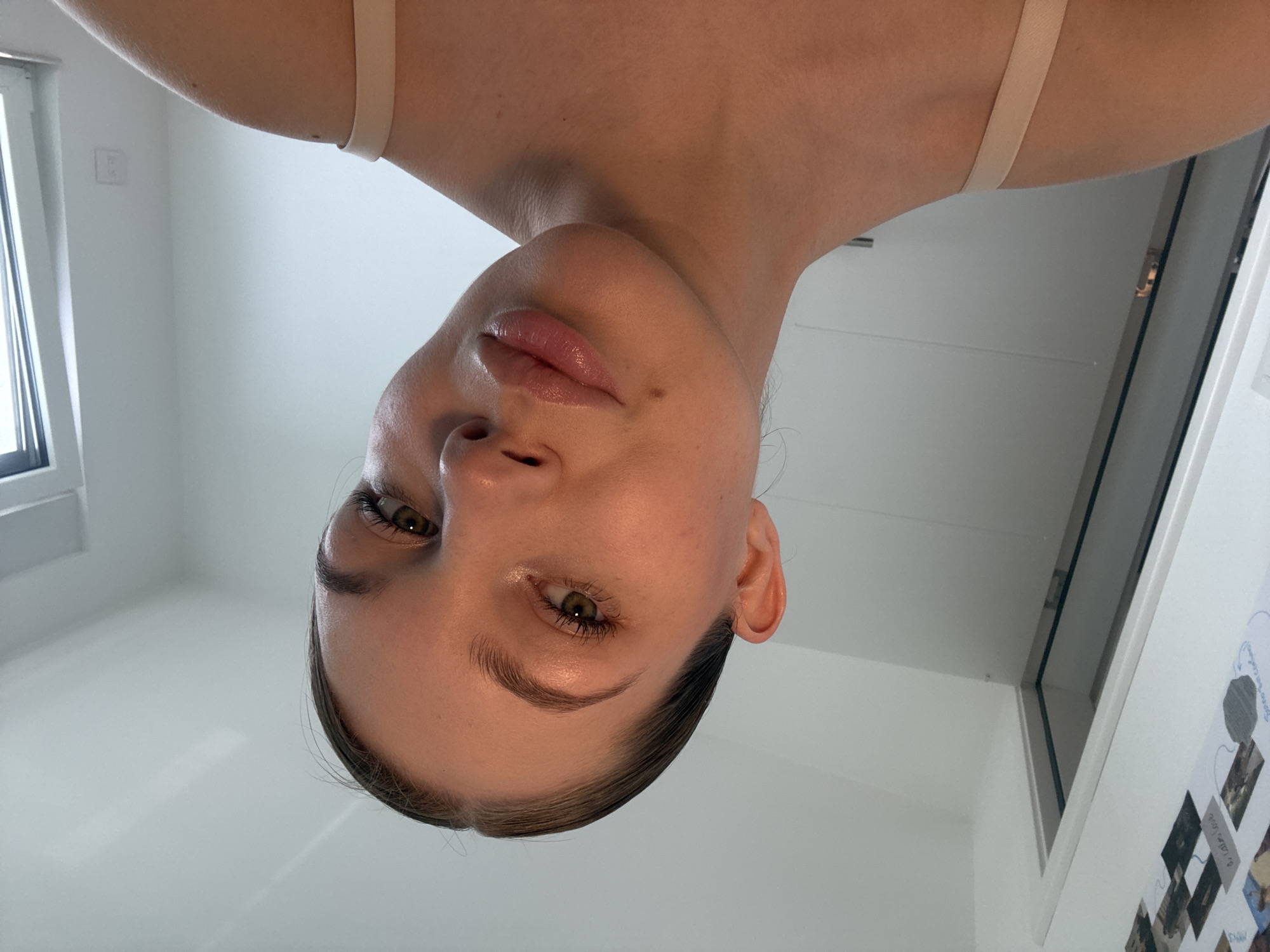A person's face is viewed from a low angle, tilted downwards. The person has fair skin, green eyes, and light brown hair pulled back. They have full lips with a hint of gloss. The background consists of a white ceiling, a window on the left, and a door or wall with some mounted pictures on the right. The scene appears to be indoors in a well-lit room. The exact location in Vozdovac Urban Municipality, Serbia cannot be determined from the image.