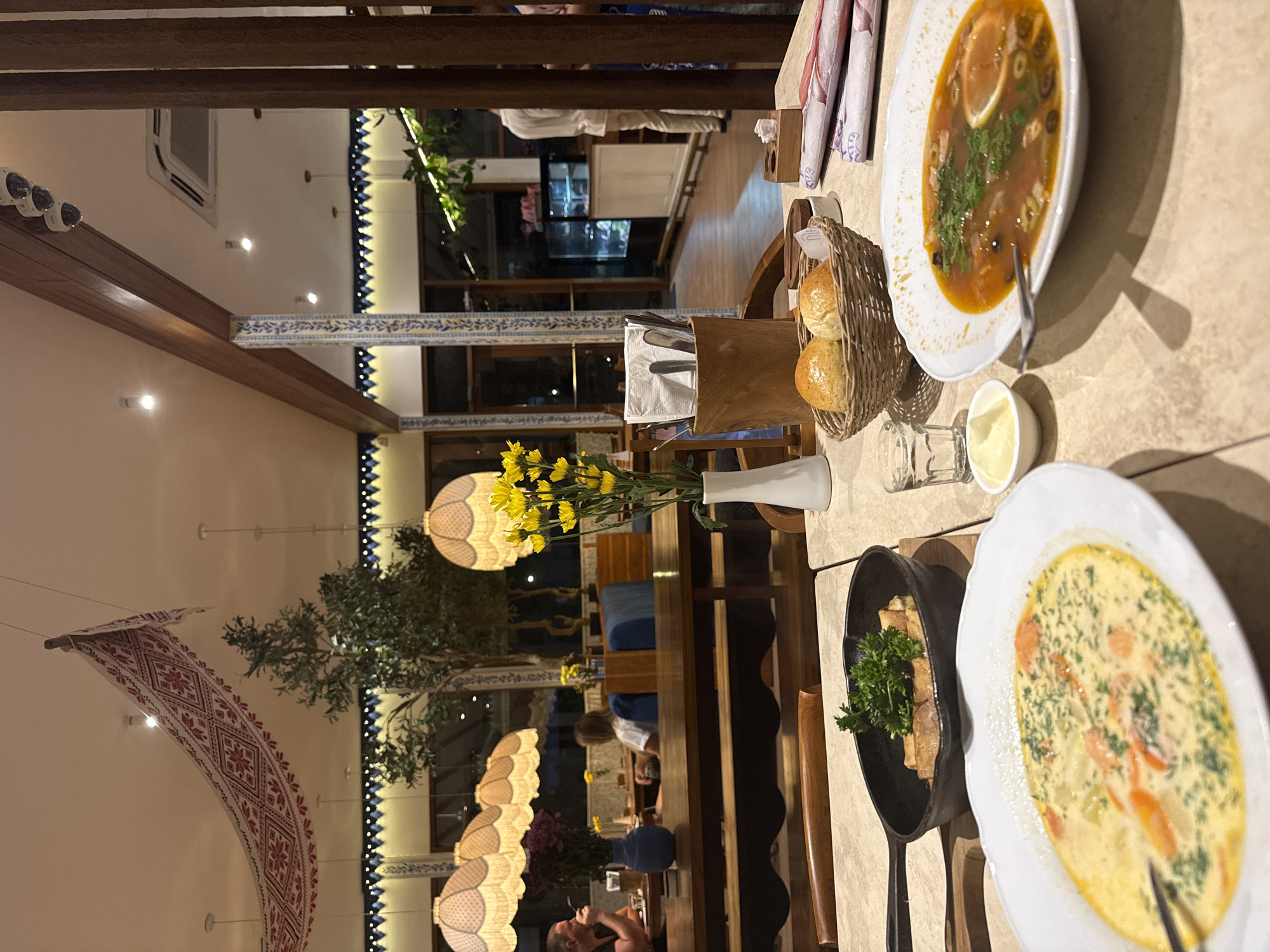 The image depicts a dining scene, likely indoors in a restaurant setting. The setting appears to be in North Kuta, Indonesia.
The primary focus is on the food laid out on the table. Two bowls of soup are visible. One is a tomato-based soup with olives and a lemon slice, while the other is a creamy soup with dill and carrot pieces. A small dish of sour cream accompanies the soup. A cast iron pan contains what appears to be wrapped food with a sprig of parsley. A basket of buns is also present.
Flowers in a vase adorn the center of the table.
The background of the restaurant includes a window with a view of trees, as well as interior decorations such as patterned lampshades and a patterned banner hanging from the ceiling. Several people are seated at other tables in the background.