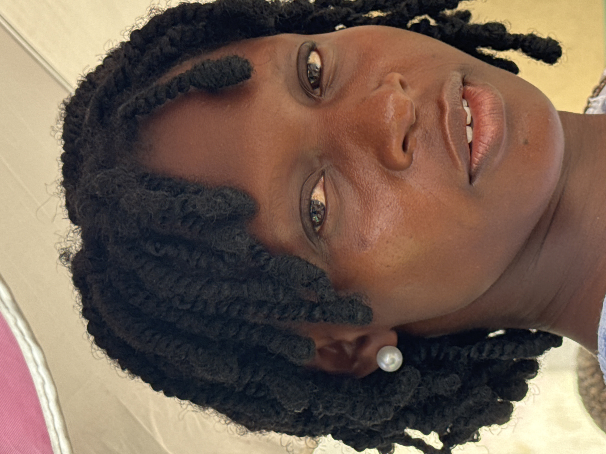 The image shows a close-up, upward-angled view of a Black woman with dark, twisted, textured hair framing her face. She wears a single pearl earring in her left ear. Her skin is smooth and has a slight sheen. Her eyes are open, and her lips are slightly parted, revealing a glimpse of her teeth. The background is indistinct but appears to be a partially visible umbrella or canopy with a pink and white section. The image was taken in Ado, Nigeria.