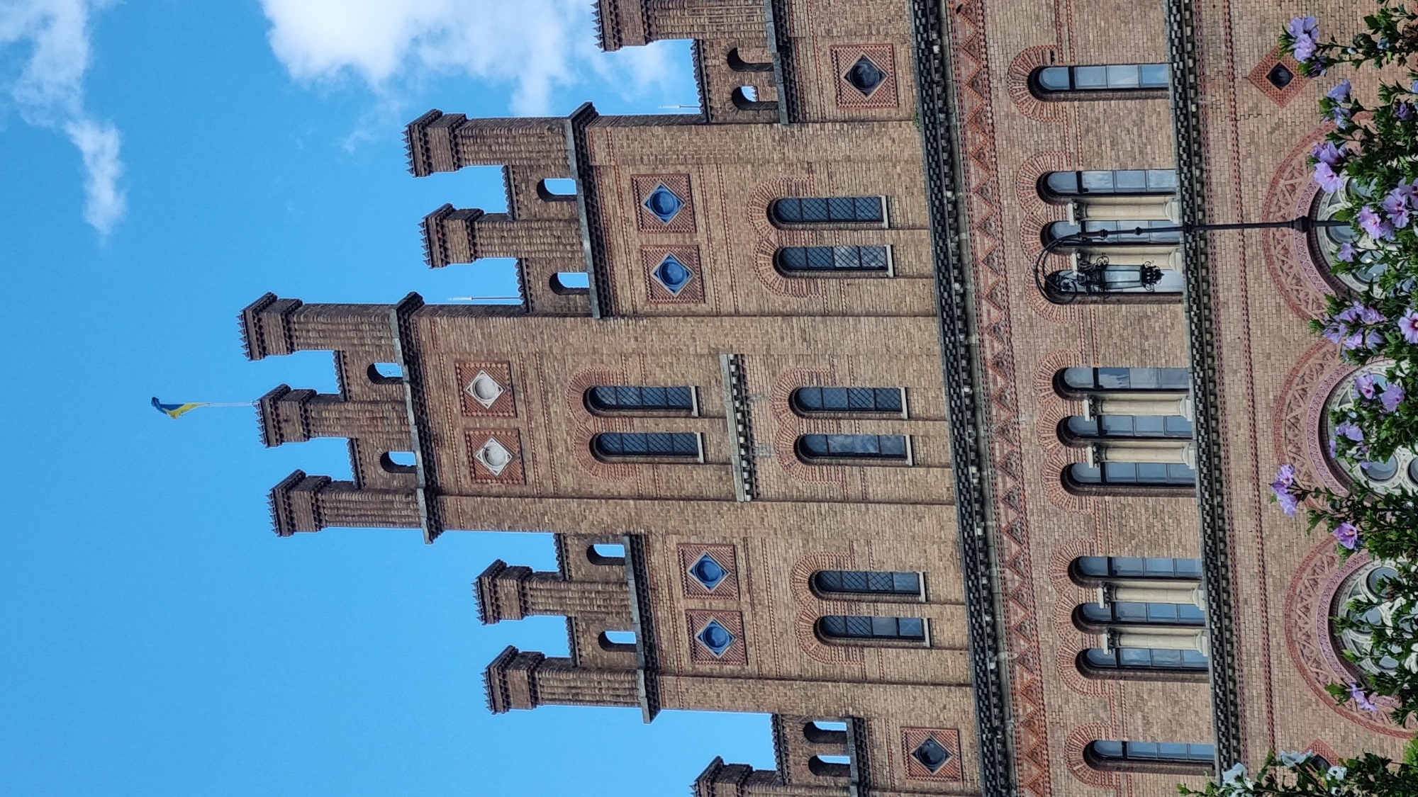 The image captures a detailed view of the central building of Chernivtsi National University, formerly the Residence of the Metropolitan of Bukovina and Dalmatia, located in Chernivtsi, Ukraine. The architectural style is a mix of Neo-Byzantine and Romanesque Revival. The building is constructed of brick with decorative elements, including patterned brickwork, diamond-shaped blue inserts, and arched windows. On the topmost part of the building, a Ukrainian flag flies against a clear blue sky with scattered white clouds. In the foreground, the upper branches of flowering bushes with purple blossoms are visible, framing the lower portion of the university building. The scene is a sunny day, highlighting the intricate details of the historic architecture.